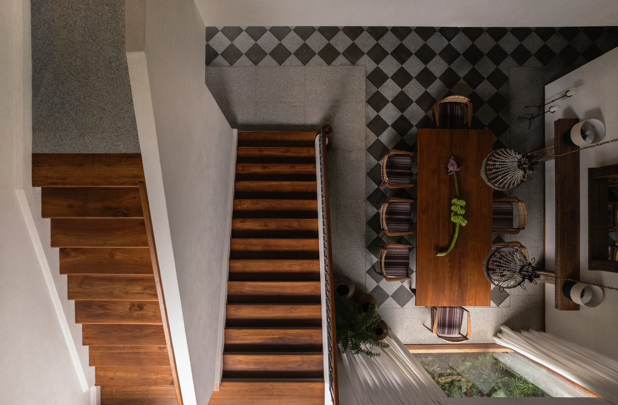Top-down view of a timber staircase beside a dining table and chandeliers on a checkerboard floor, with a planted well below