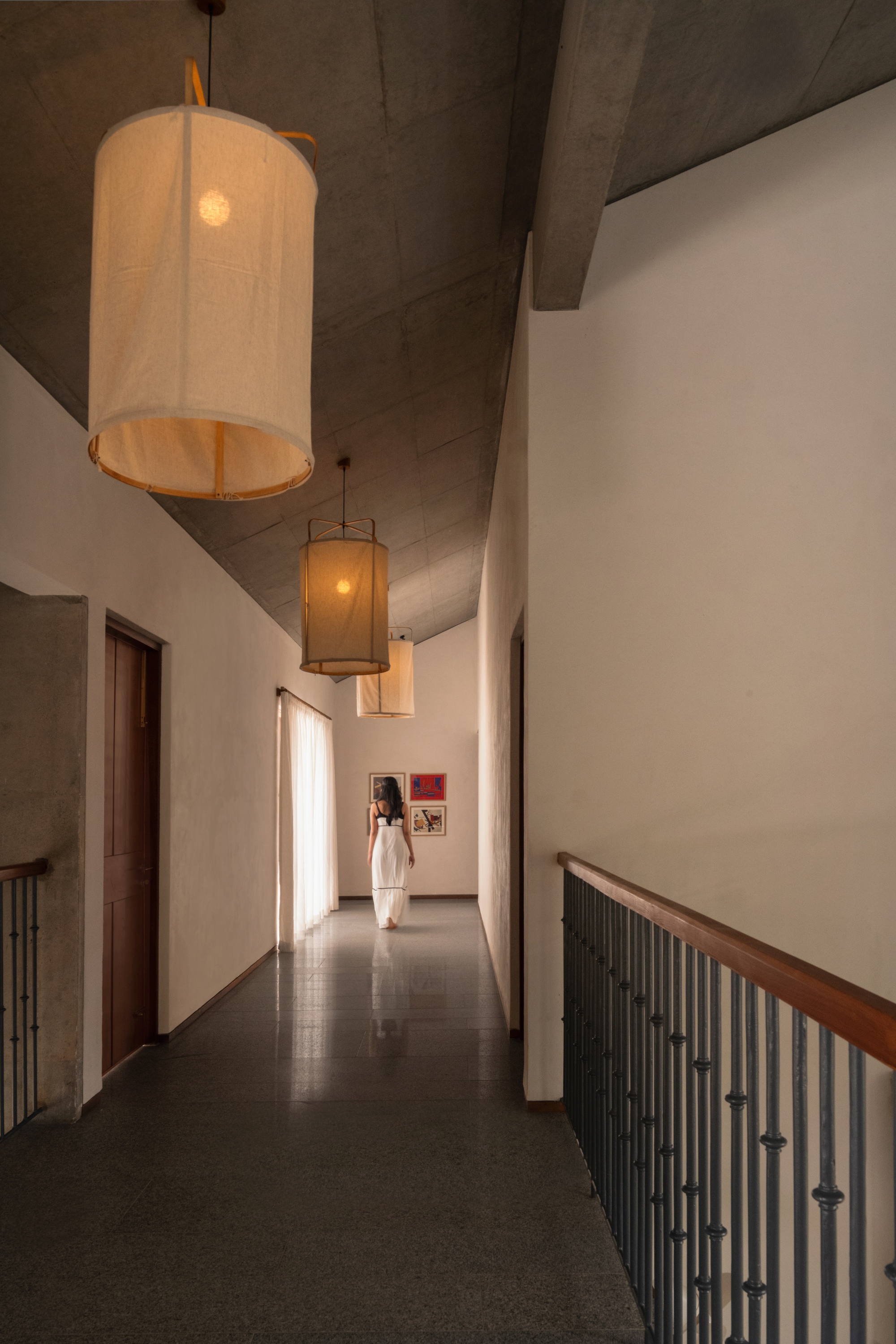 Upper corridor with fabric pendant lamps, a concrete vaulted ceiling and a timber-topped railing, a woman in white walking toward framed art