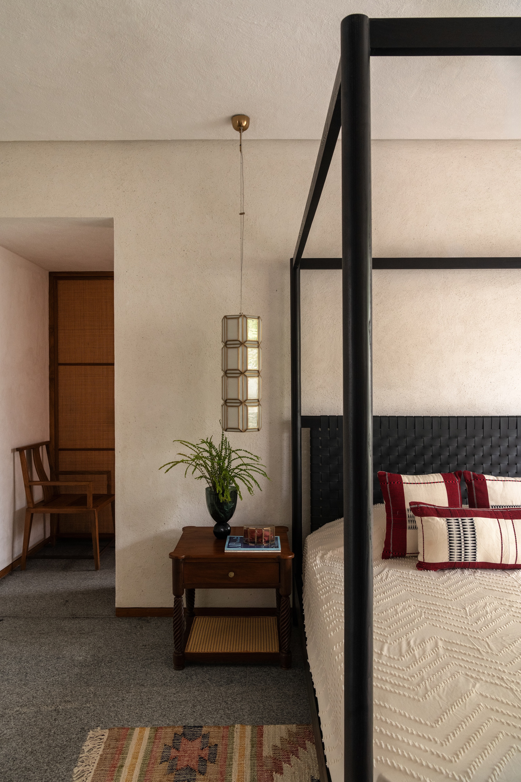 Bedside corner with a dark wood side table, fern in a vase and a brass lantern pendant beside a black four-poster bed