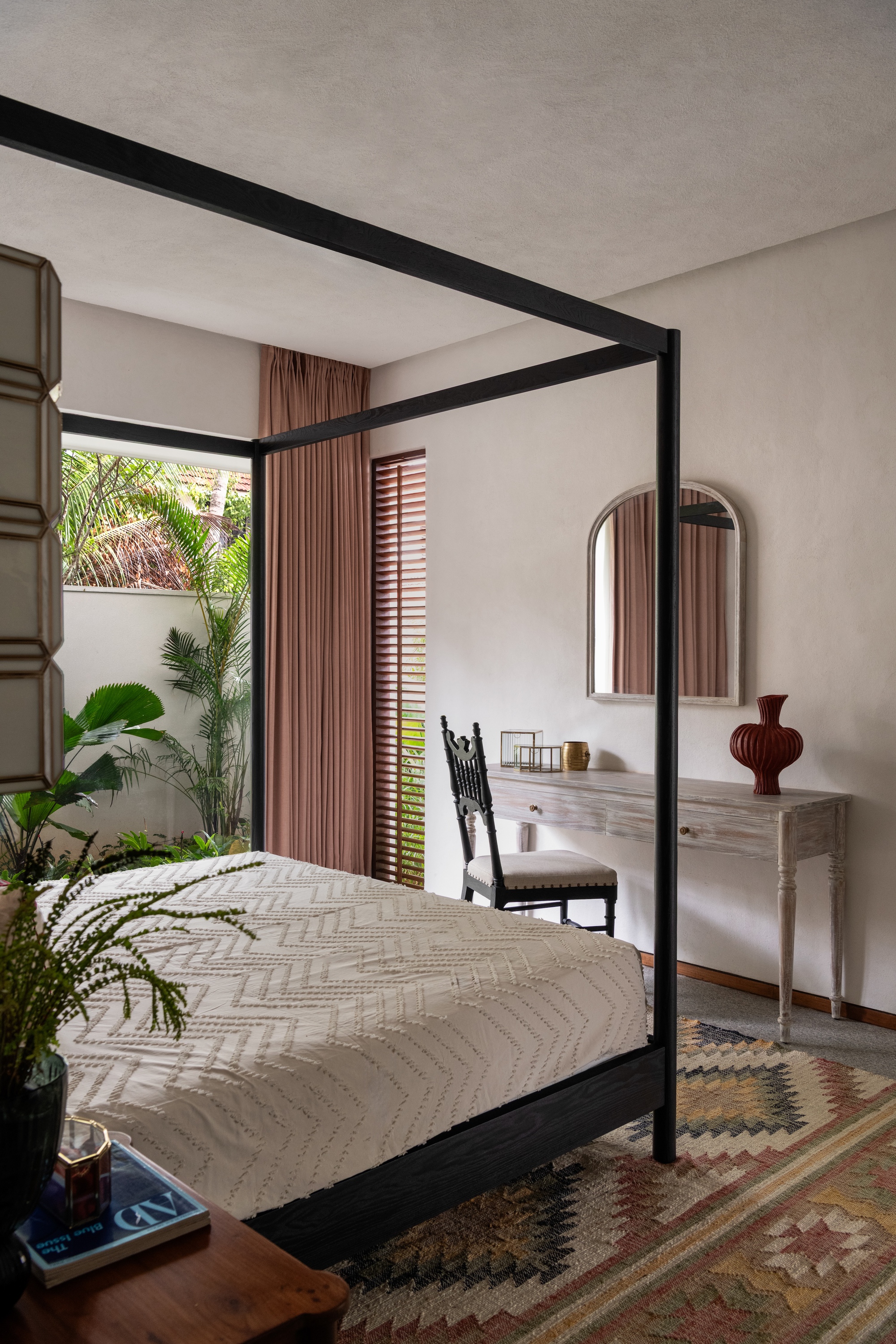Bedroom with a black four-poster bed, kilim rug and curtained window opening onto a green garden courtyard in daylight