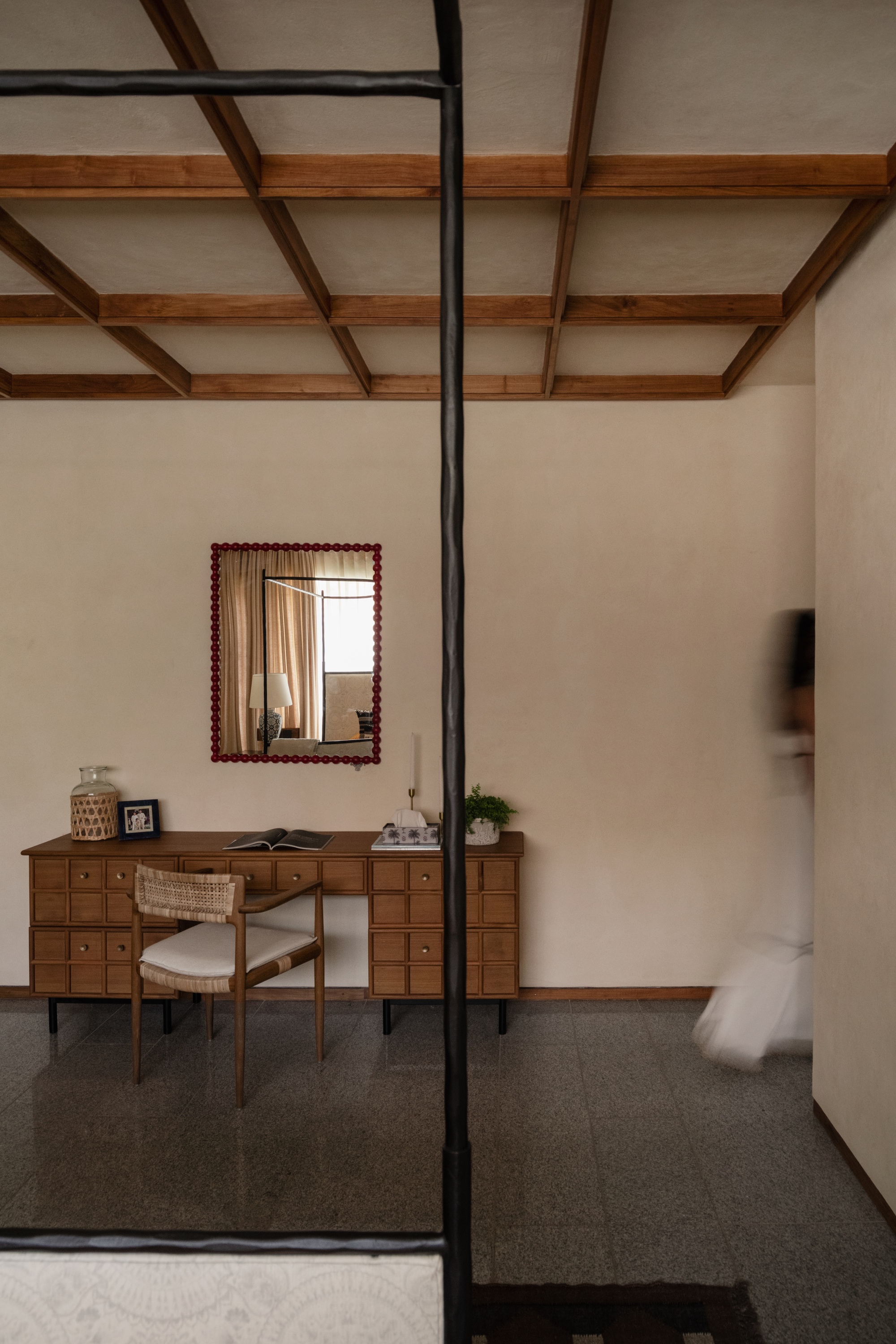 Wooden desk with small drawers and a beaded red mirror under an exposed timber beam ceiling, a blurred figure passing at right