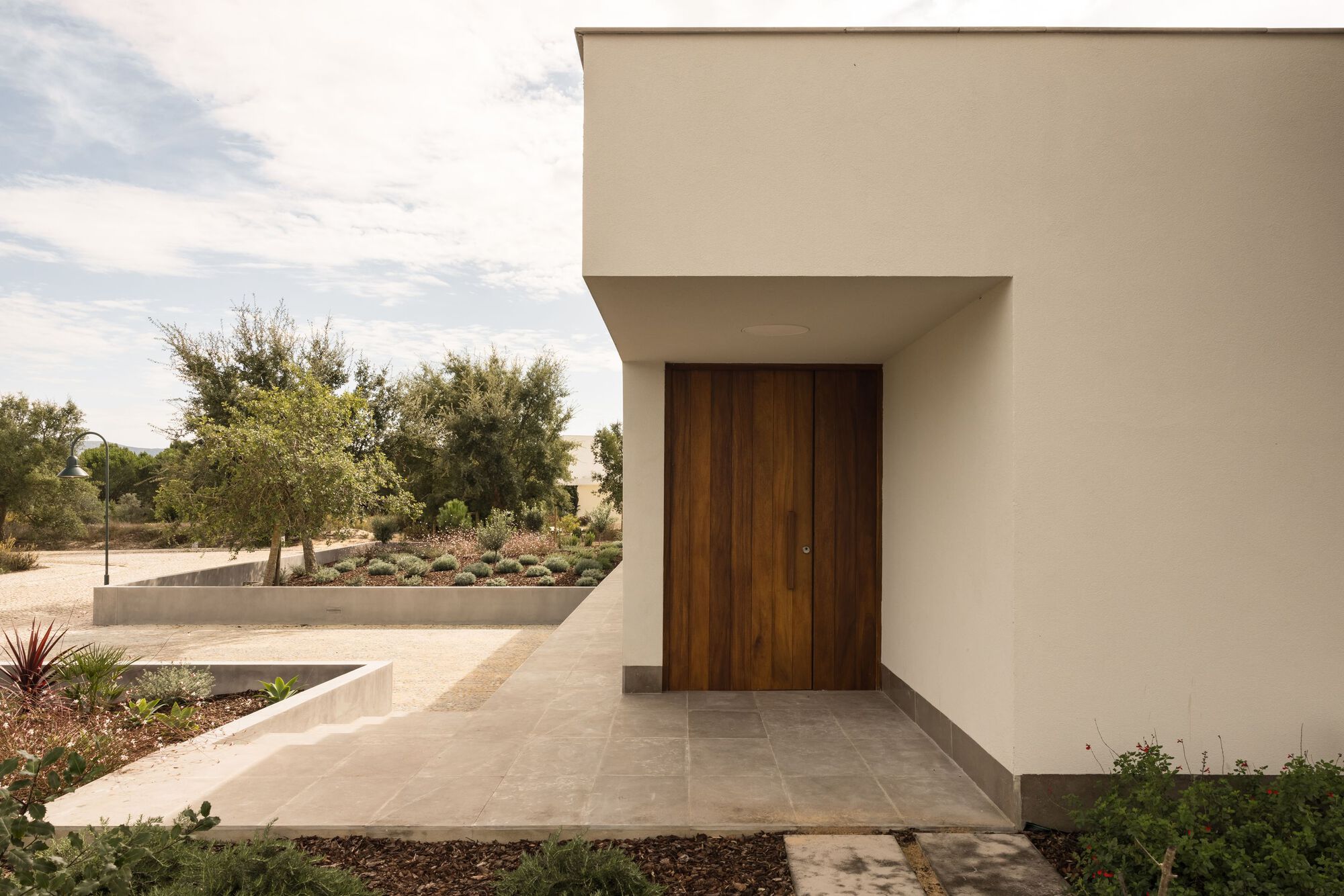 Timber entrance door recessed beneath a white overhang, with a stone-paved path and planted gravel beds in soft afternoon light
