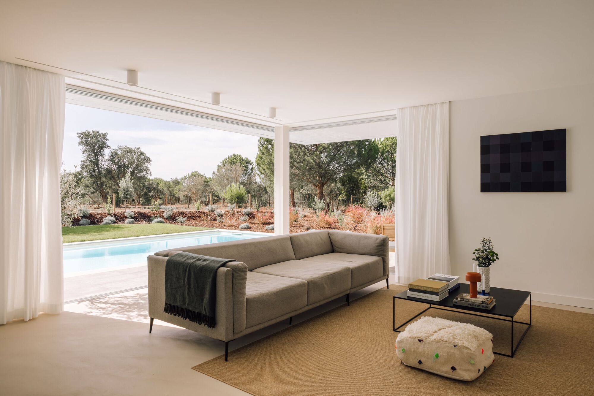 Living room with a grey sofa and black coffee table, sheer curtains framing a sliding glass opening to the pool and pines