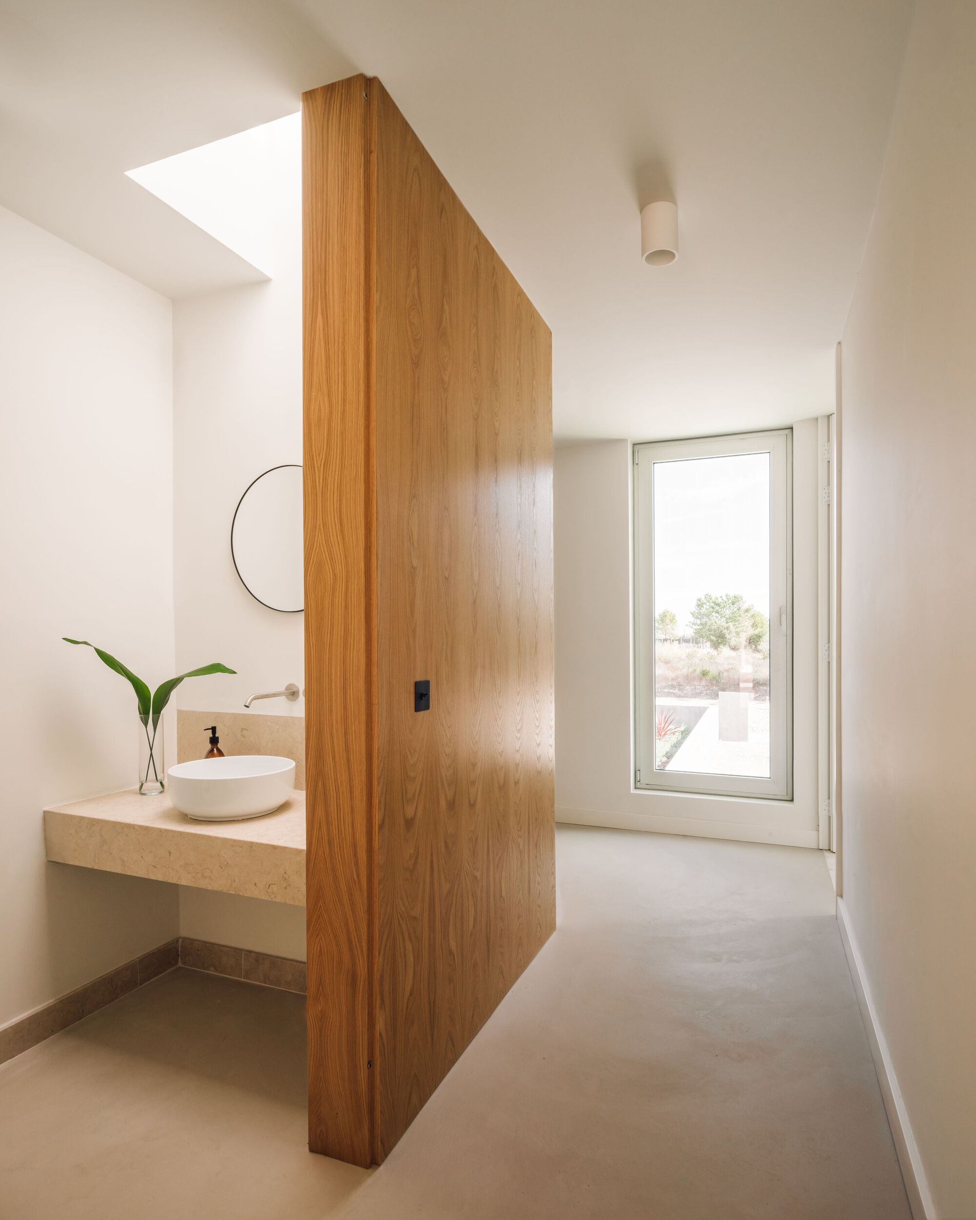 Oak partition wall screening a stone washbasin with a round mirror, a skylight above and a tall window at the corridor end