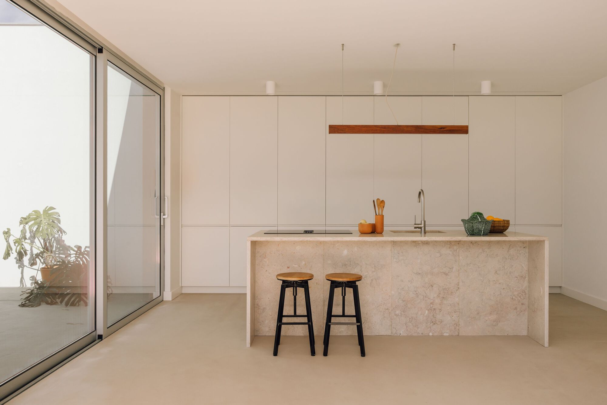 Kitchen with a stone-clad island, two black wooden stools and floor-to-ceiling white cabinets beside a sliding glass wall opening to a courtyard plant