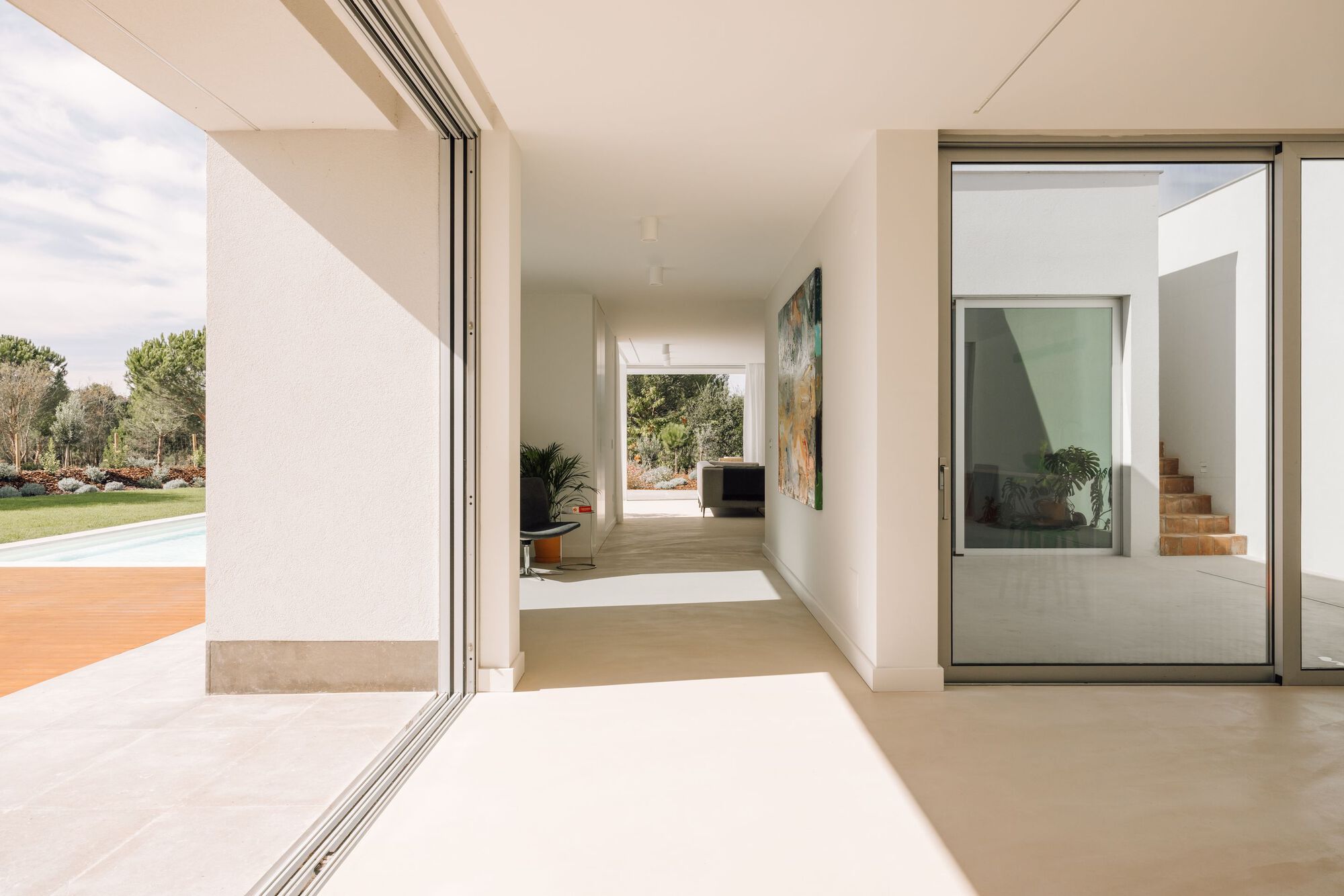 Glazed corridor with sliding doors opening to a pool terrace, a painting on the wall and a terracotta stair beyond the glass