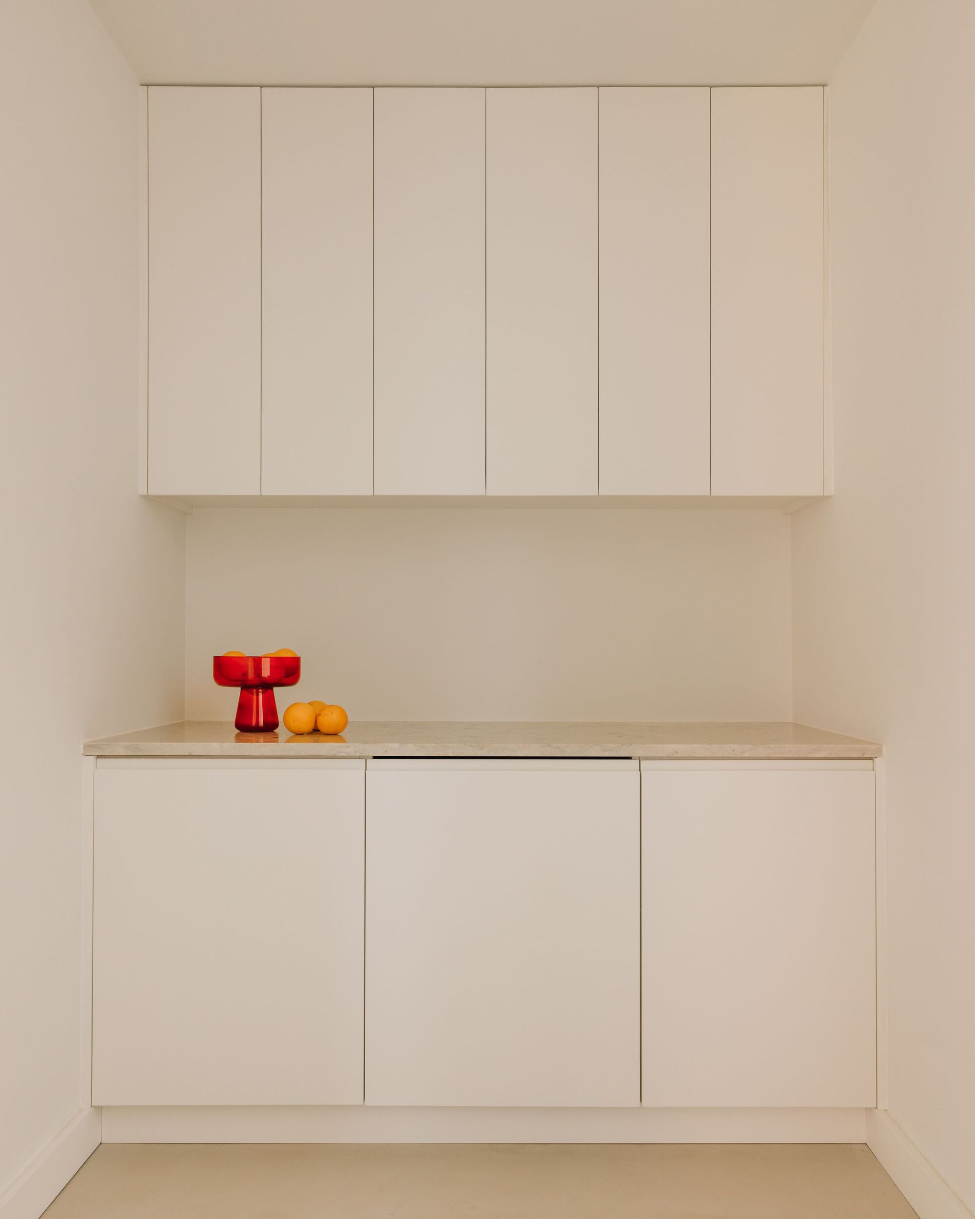 Built-in white cabinetry with a stone countertop holding a red glass bowl and oranges in soft daylight