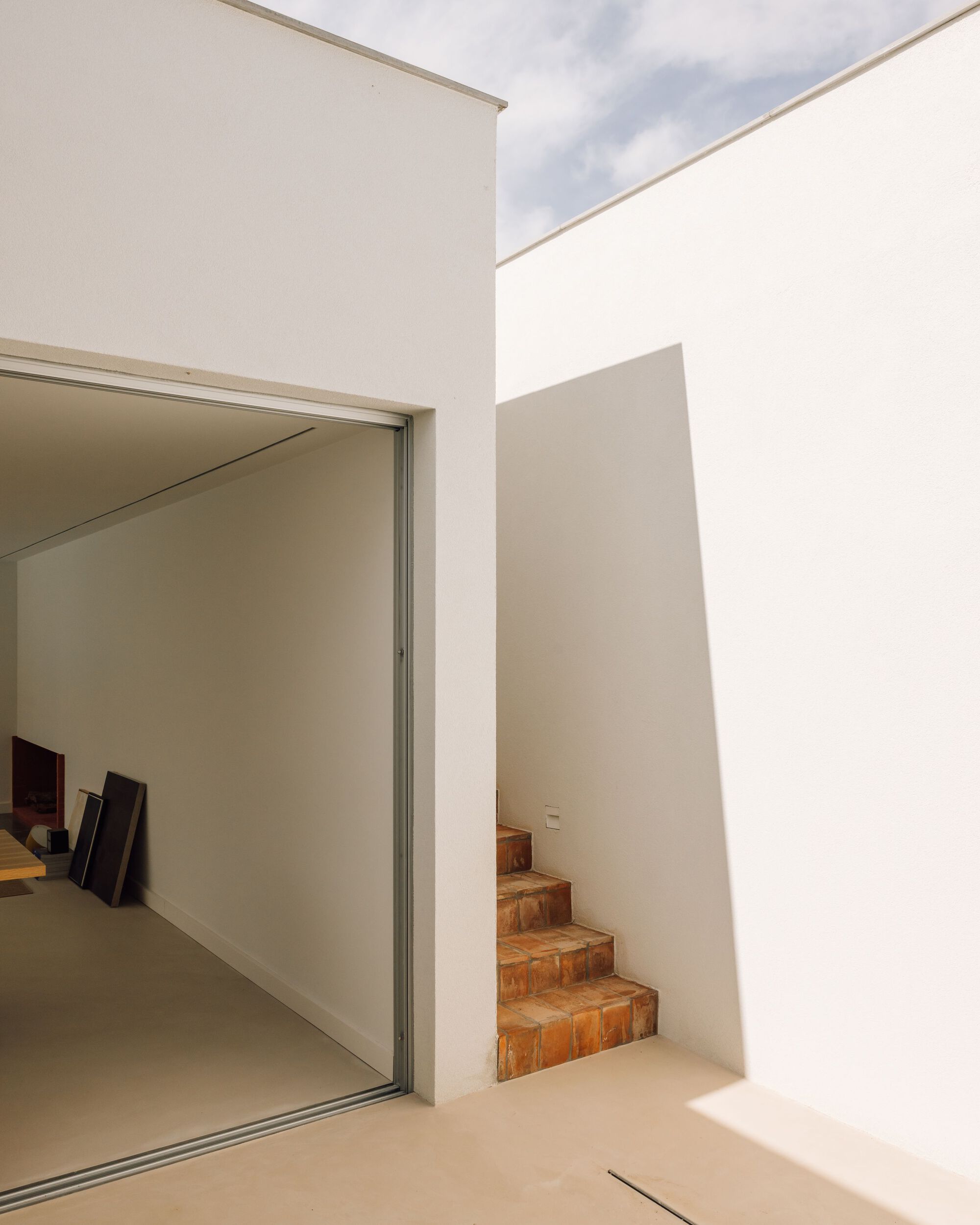 Whitewashed courtyard with a terracotta stair beside an open sliding door, sharp shadows cast under a partly cloudy sky