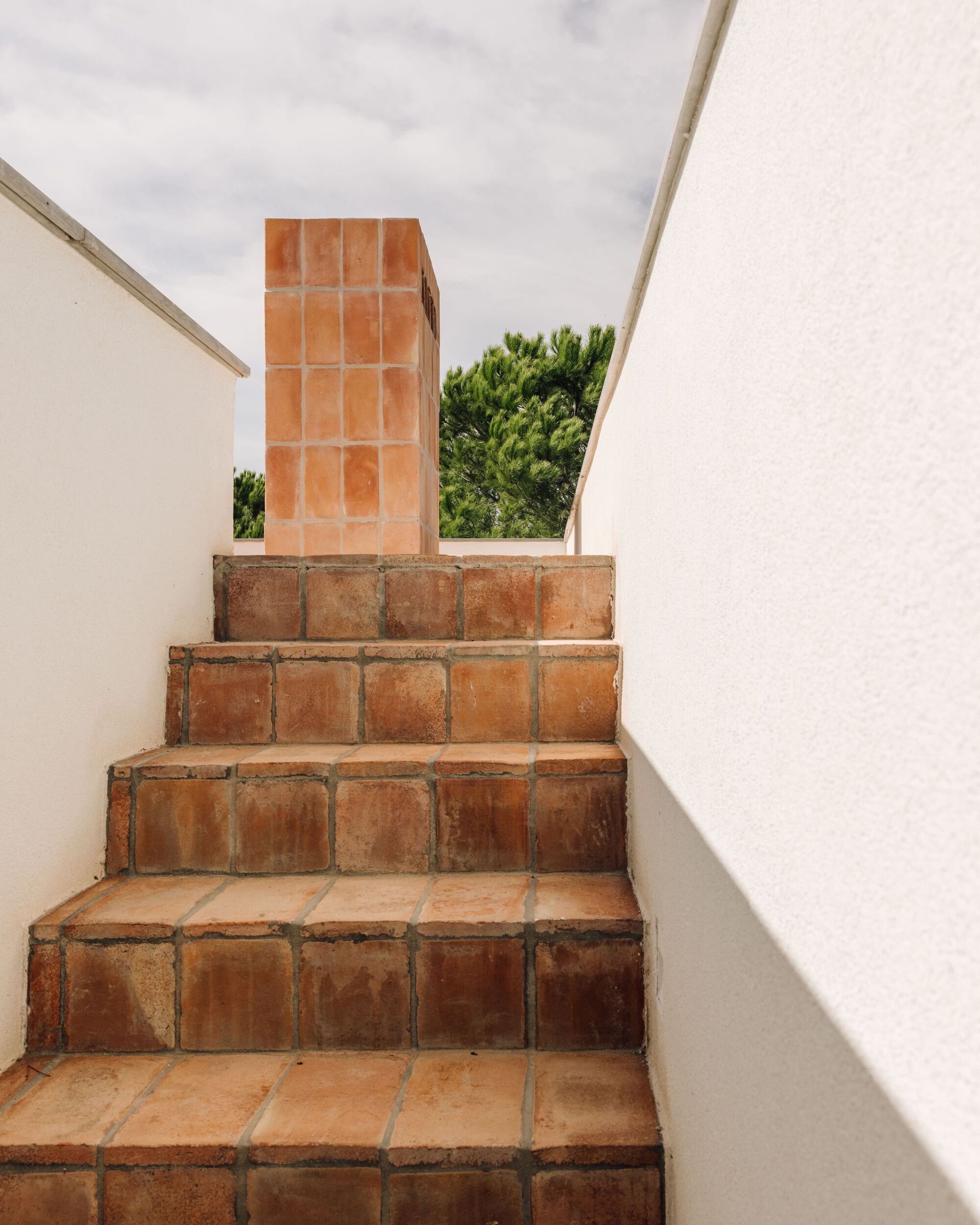 Terracotta tile steps rising between white walls toward a tiled chimney and pine trees under overcast sky
