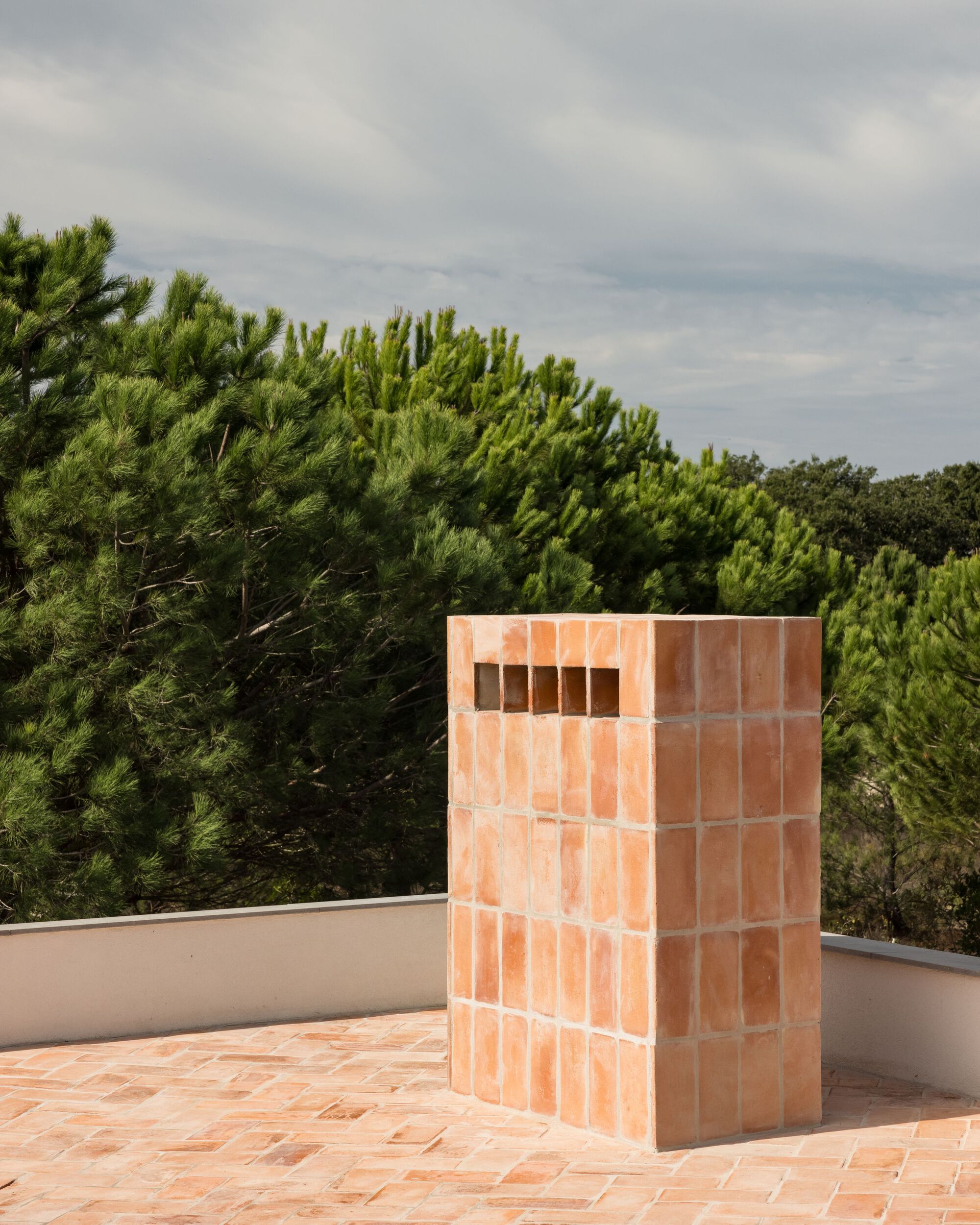 Terracotta-tiled chimney with small slot openings on a tiled roof terrace, framed by pine canopy and cloudy sky