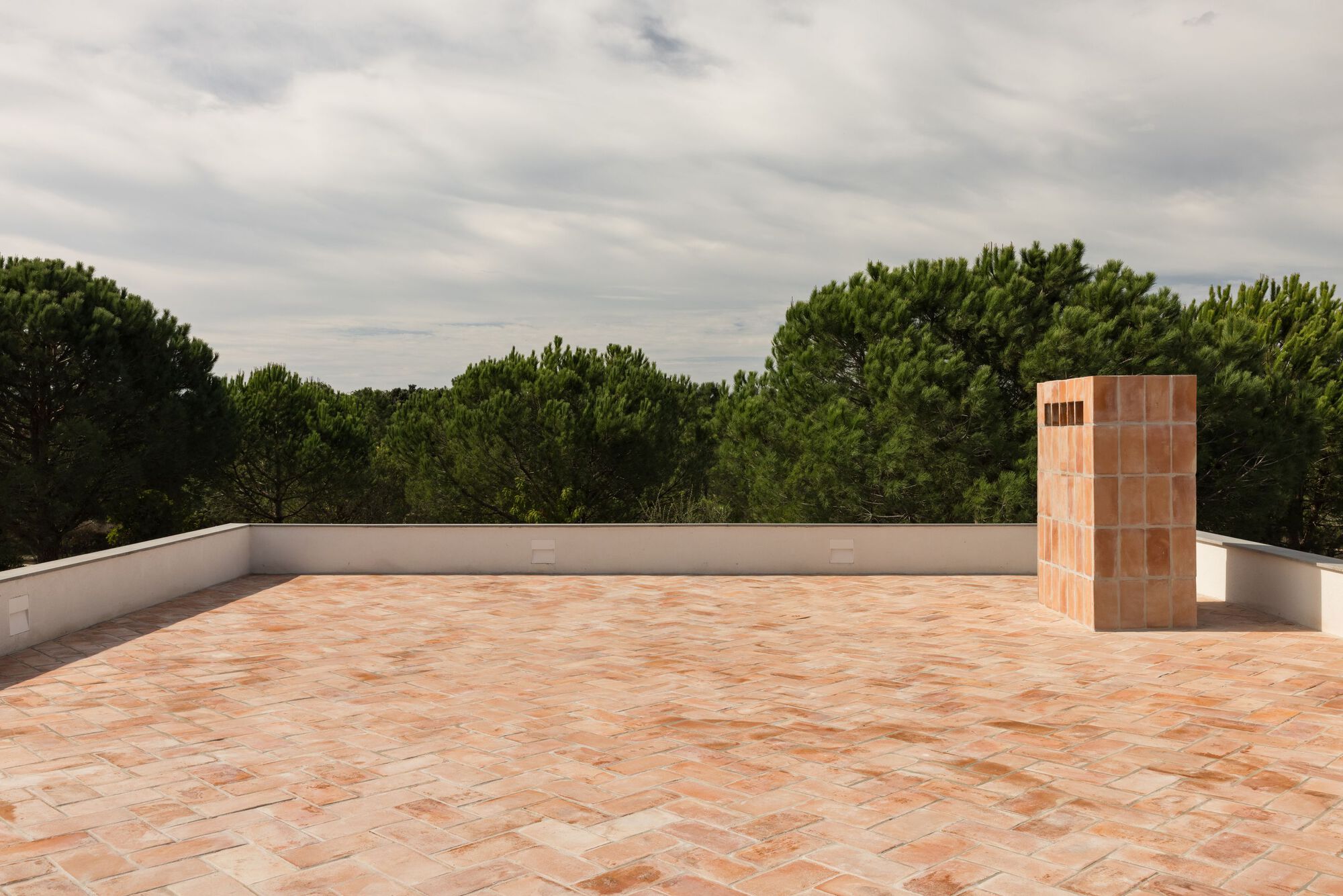 Wide roof terrace paved in terracotta tile with a tiled chimney, low white parapet and pine trees under grey clouds