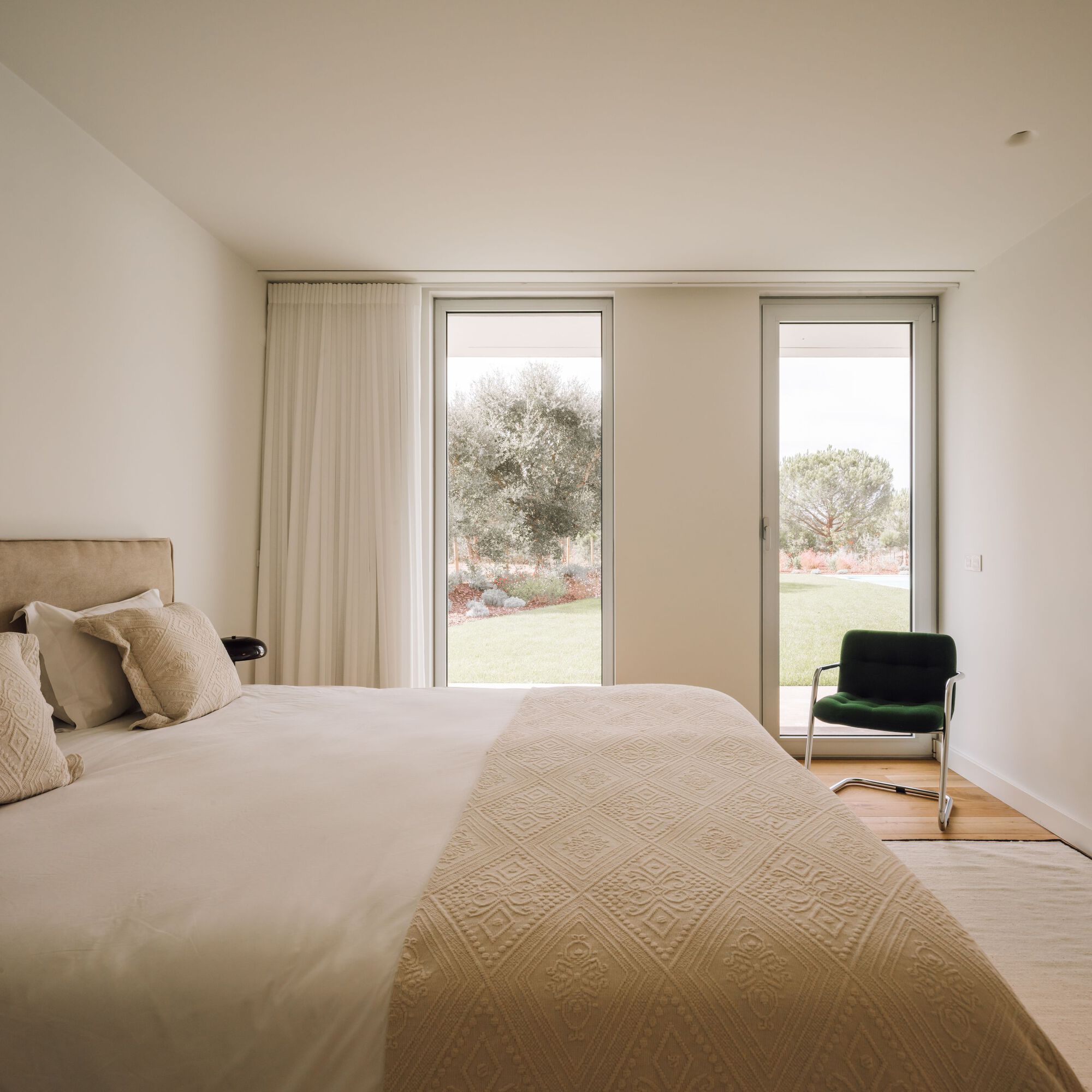 Bedroom with a quilted cream bedspread and a green cantilever chair beside glass doors opening to an olive tree and lawn