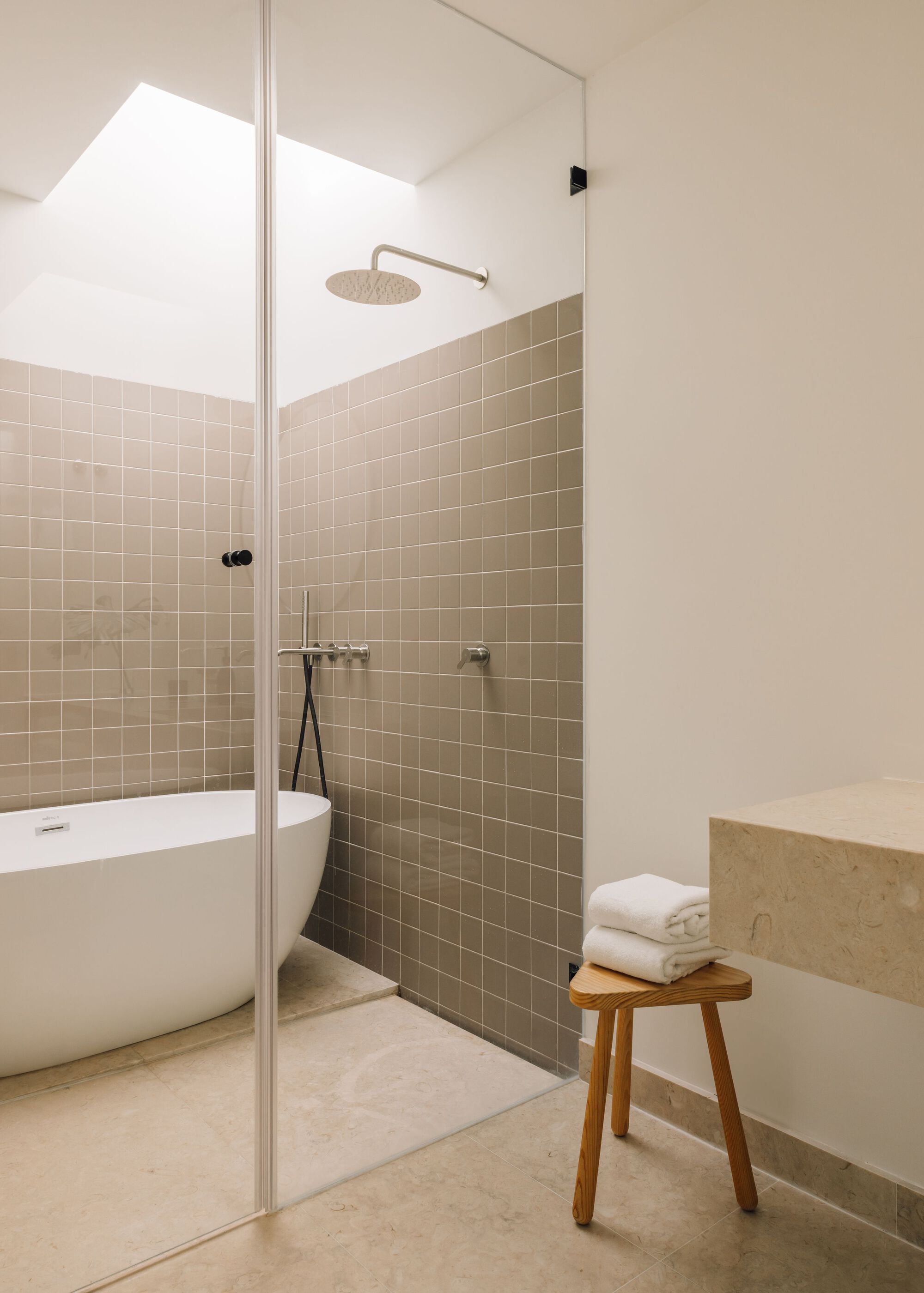 Glass-enclosed shower with taupe square tiles, a freestanding white tub and a wooden stool holding folded towels under skylight light