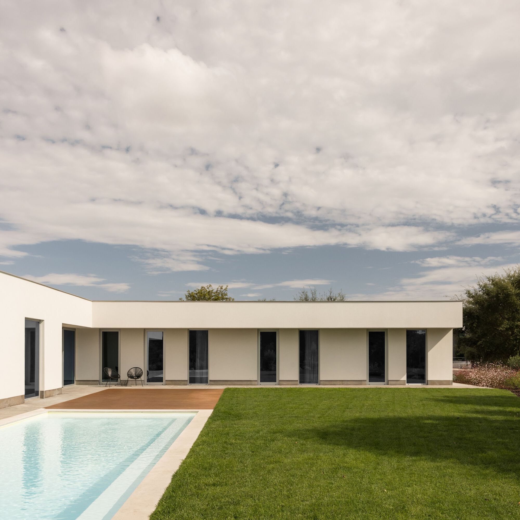 Single-storey white facade with tall glazed doors, a timber deck and a pool edge beside a green lawn under cloudy sky
