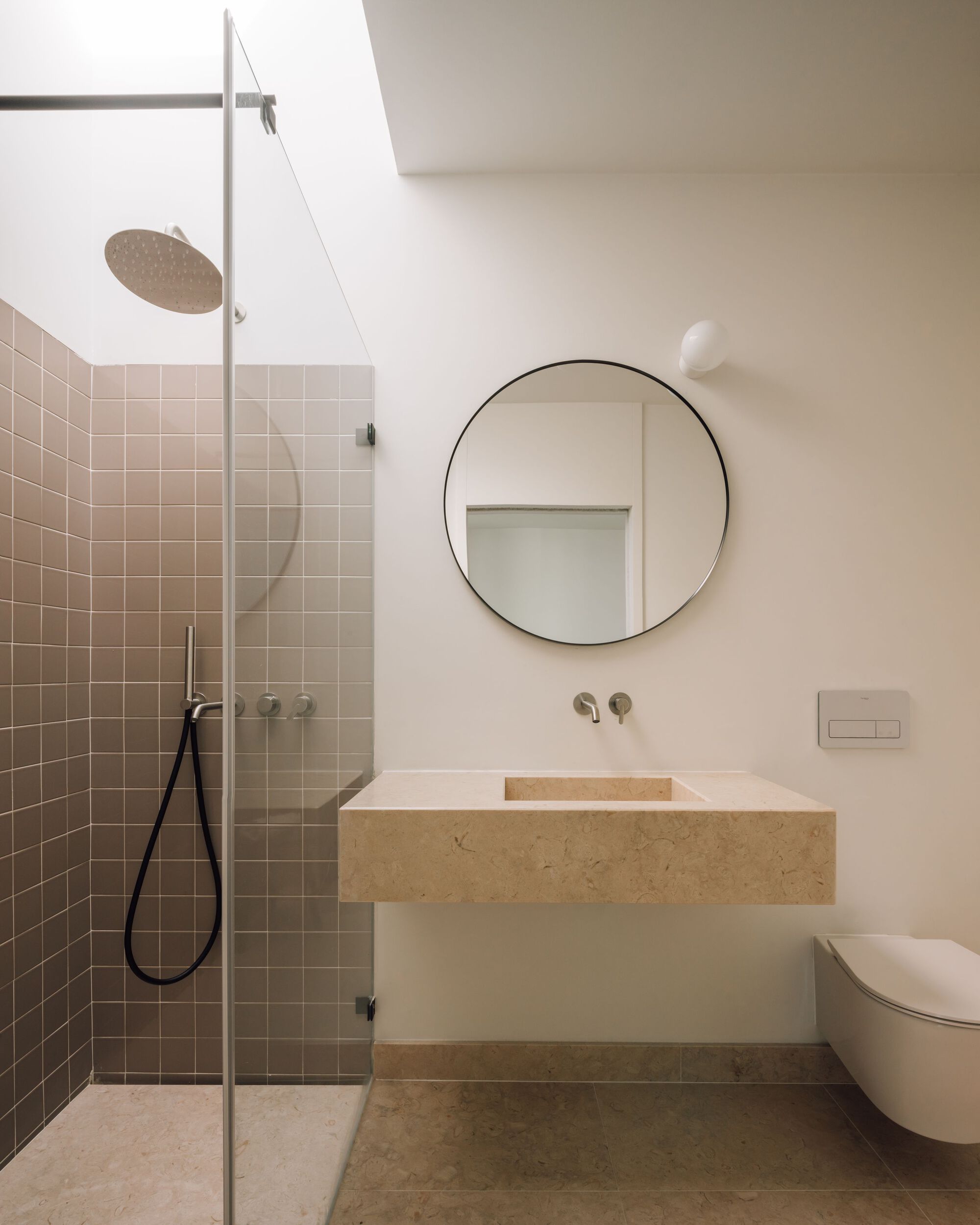 Bathroom with a floating limestone basin, round black-framed mirror and glass-enclosed shower lined in dusty pink square tiles under soft daylight