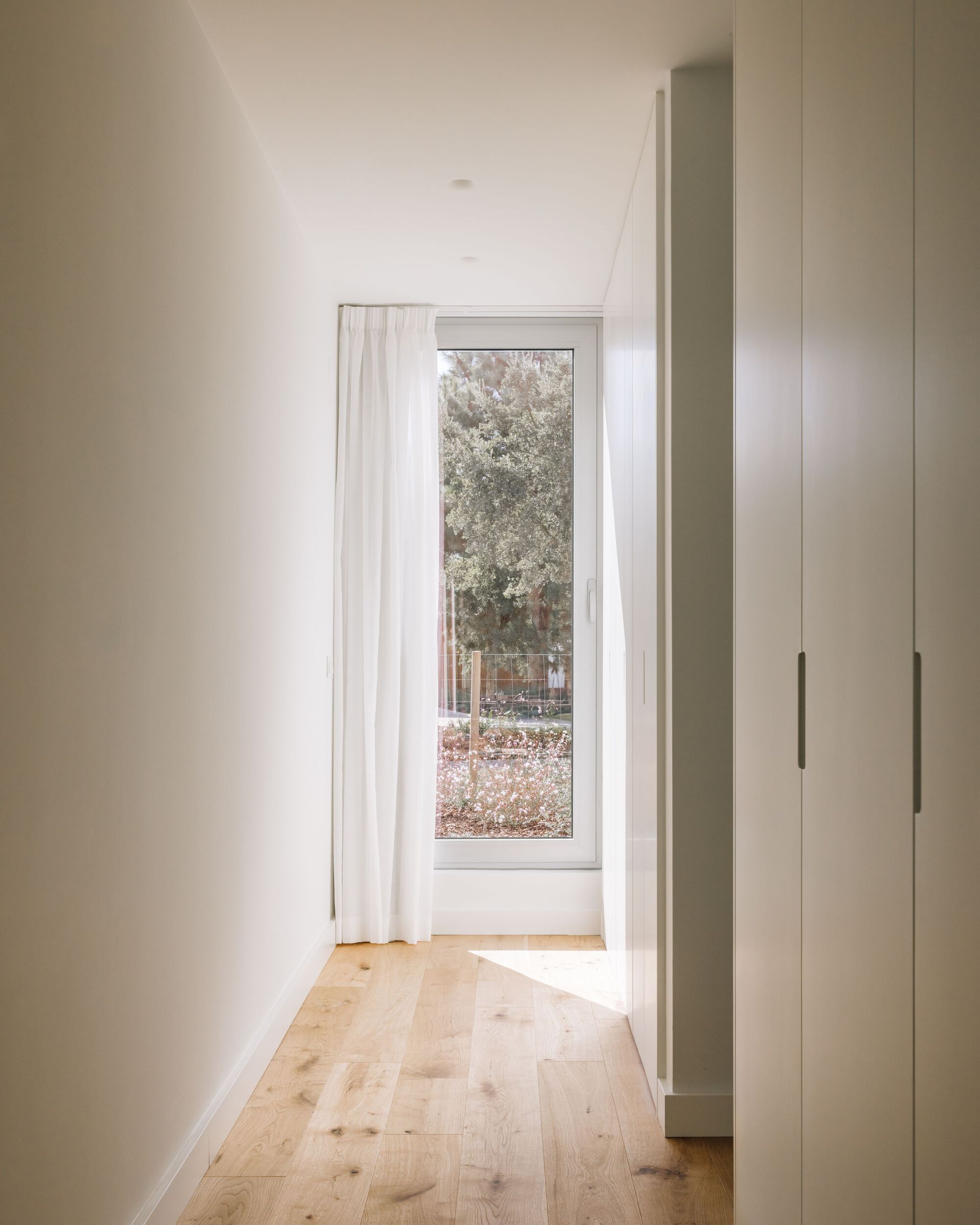 Narrow hallway with white built-in wardrobes and a sheer curtain beside a tall window, sunlight falling across oak floorboards