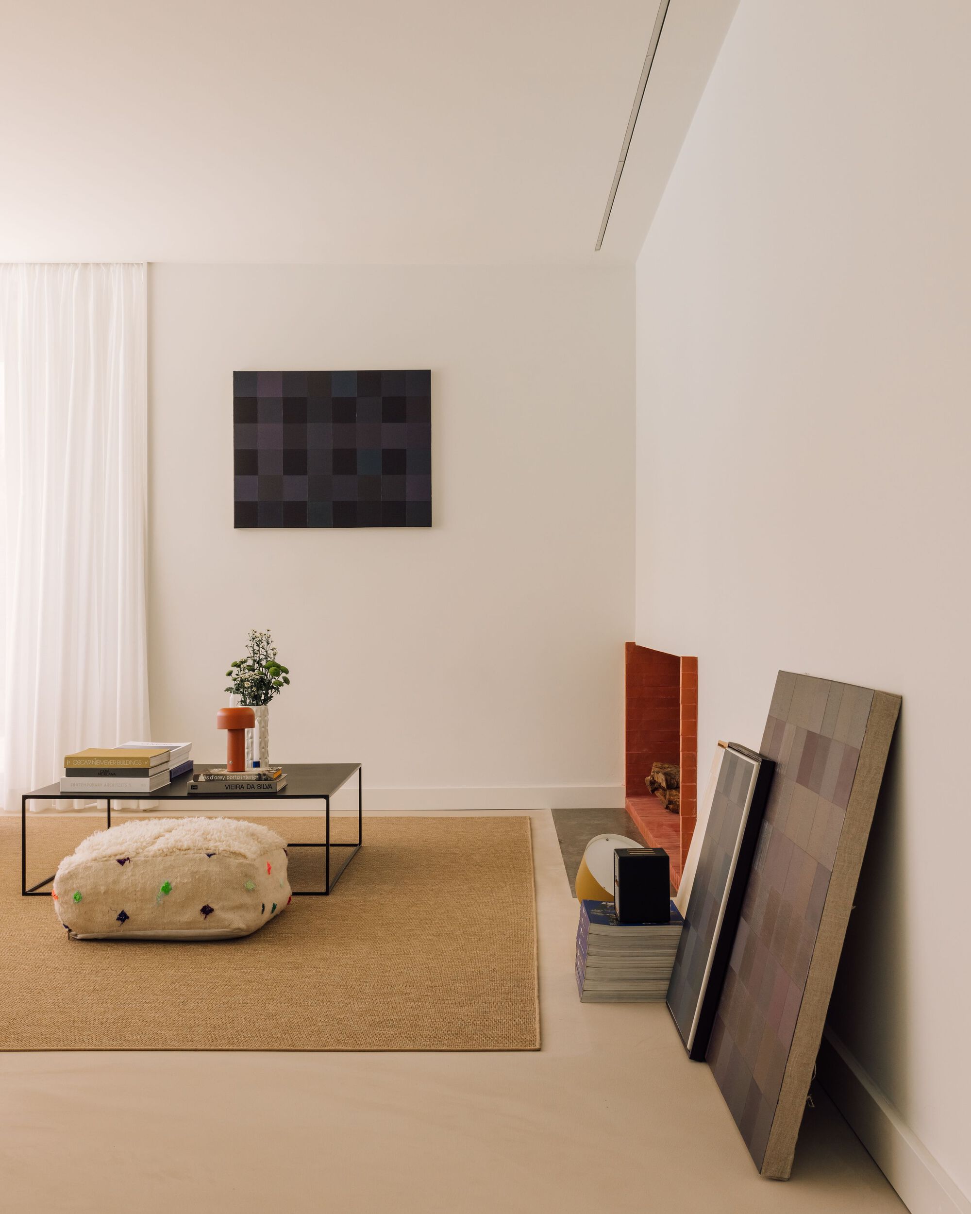 Living room corner with a low black-framed table, woven rug and pouf, checkered paintings leaning against the wall near the brick fireplace
