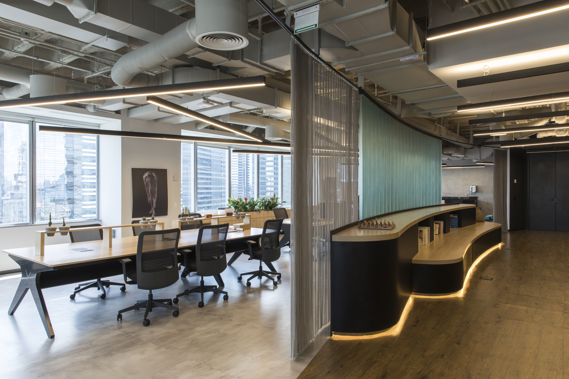 Open office with a curved black counter edged in warm under-lighting beside a mesh curtain and long timber desk, city towers in the windows