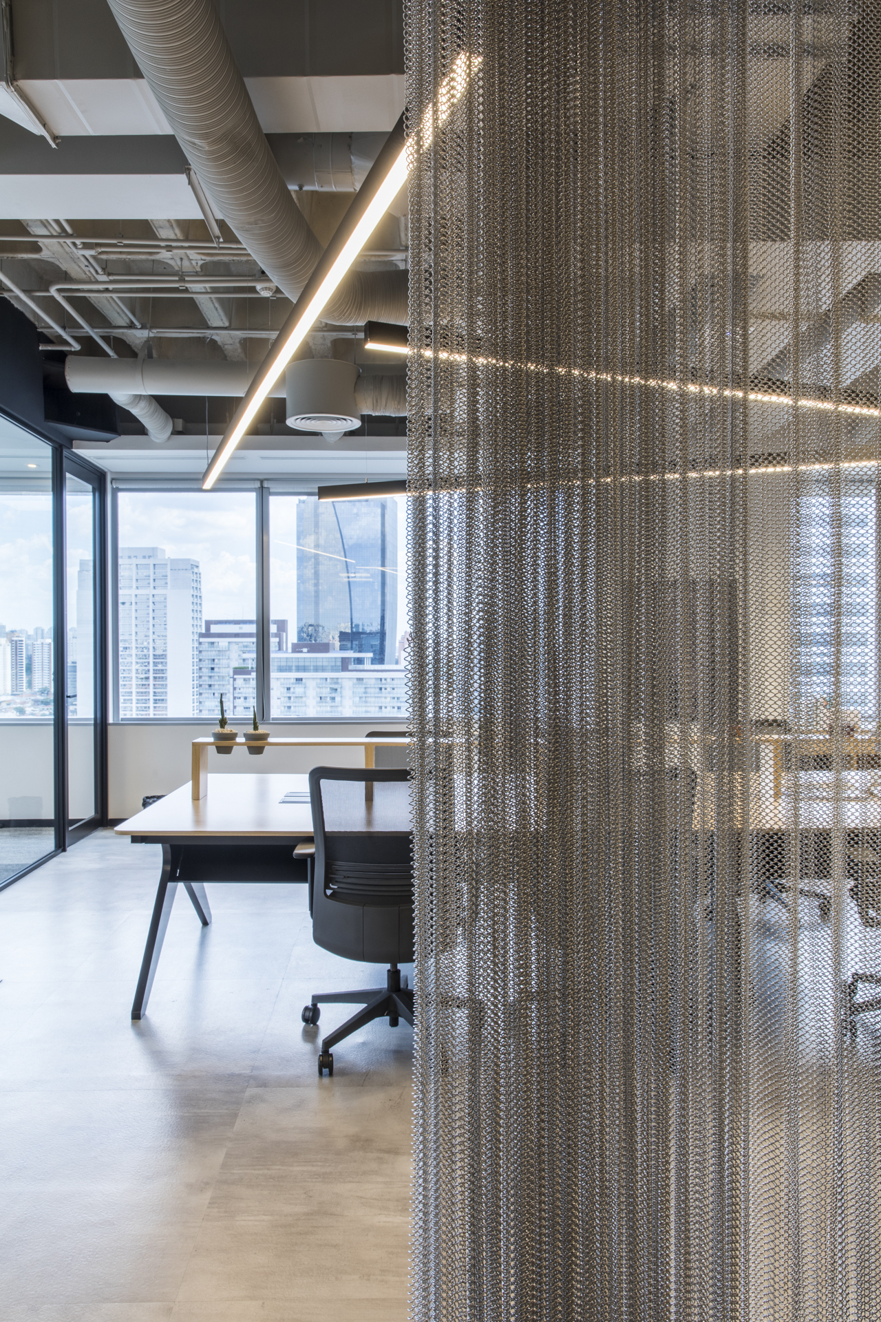 Hanging metal mesh curtain in the foreground with a desk and task chair beyond, skyline visible through daylit office windows