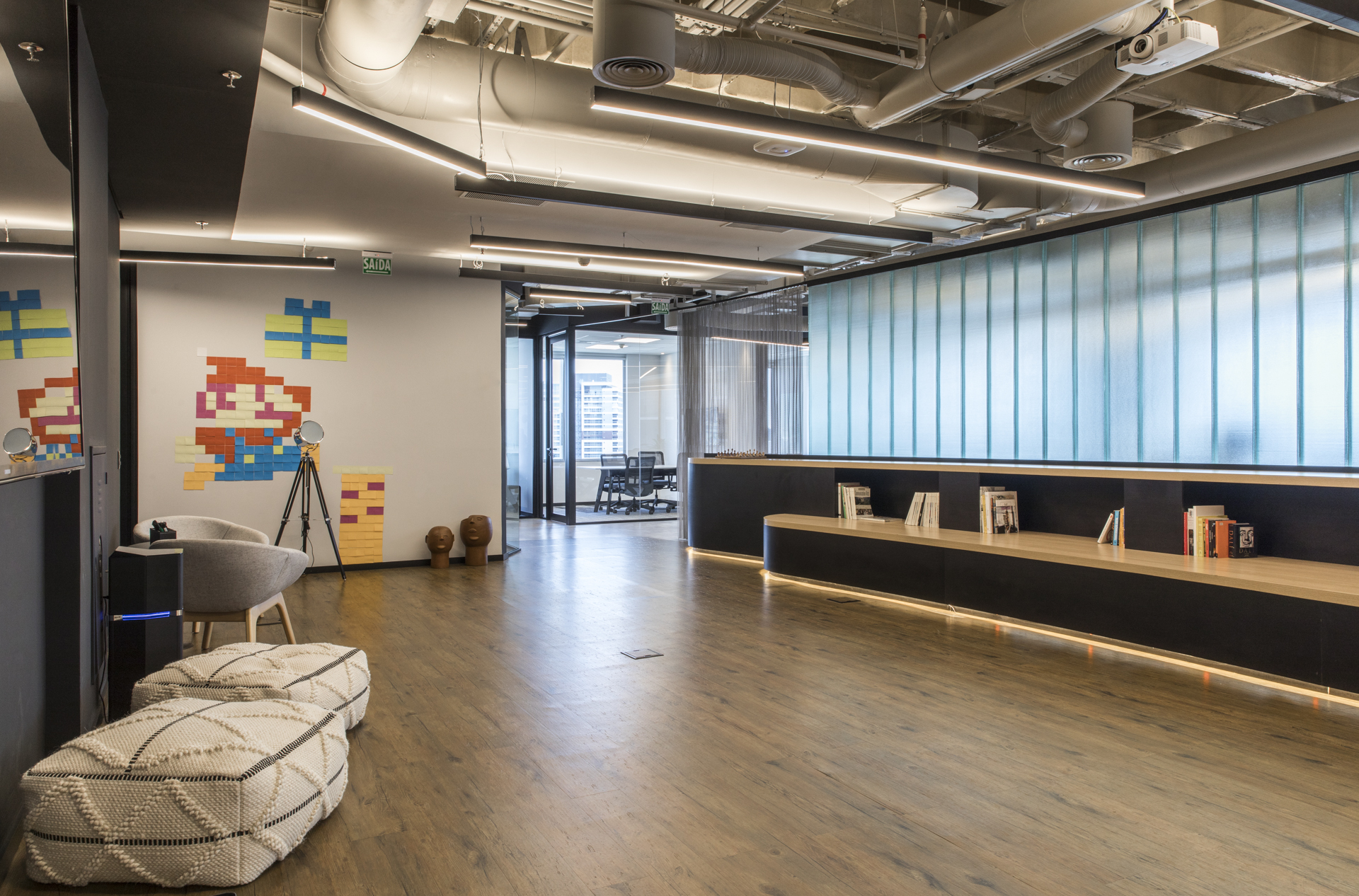 Lounge with woven pouffes, pixel-art sticky notes on the wall and a low shelf beneath translucent ribbed glass panels