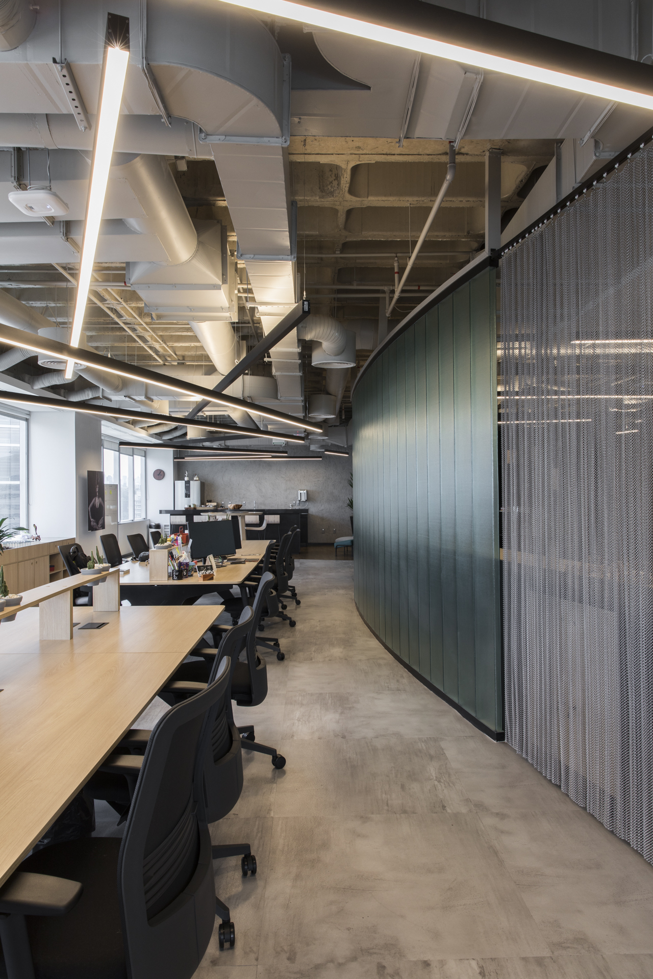 Curved green glass partition and mesh curtain running alongside a long timber desk, exposed ducts and light bars overhead