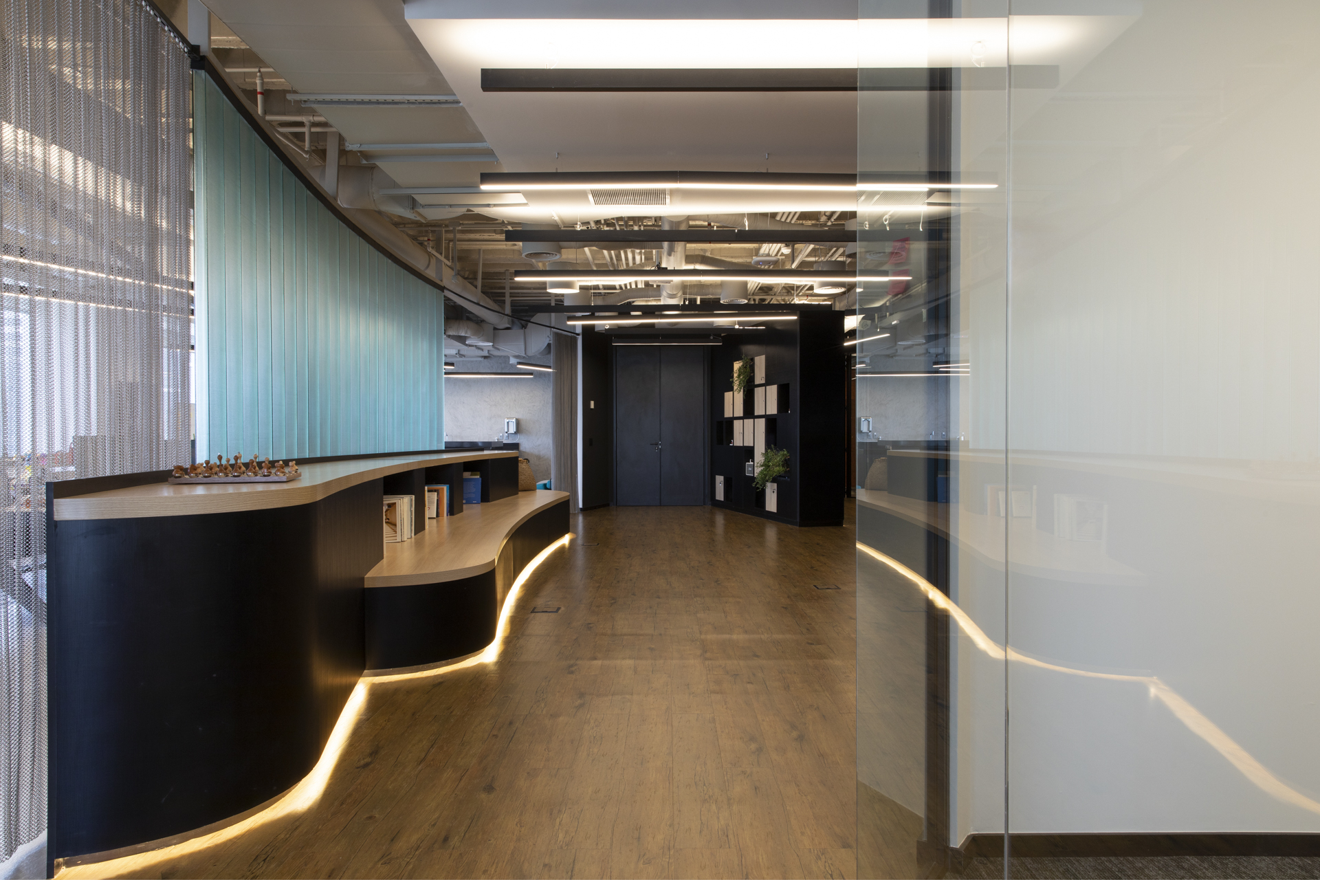 Wood-floored corridor with a curving black counter glowing along its base, mesh curtain at left and a glass pane reflecting light at right