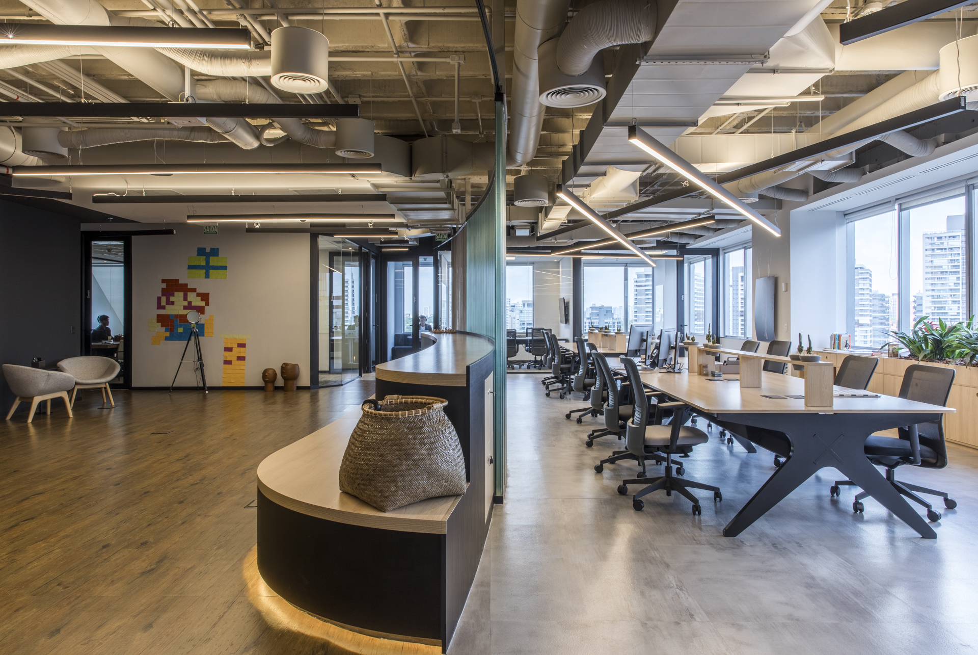 Curved wood-topped bench with a woven basket beside a long desk row, under exposed ducts and city-facing windows