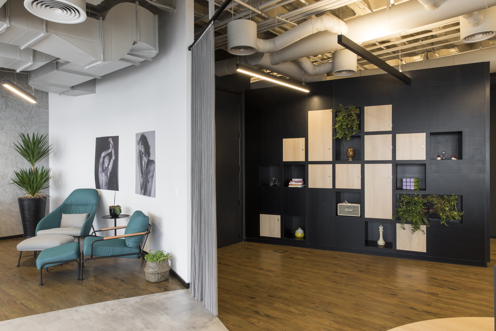 Black storage wall with pale wood doors and plant niches beside teal armchairs and a metal mesh curtain