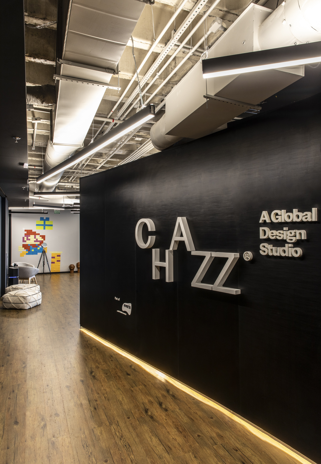Black wall with raised white lettering and a warm strip light along the floor, beside a corridor of exposed ducts and pixel-art mural