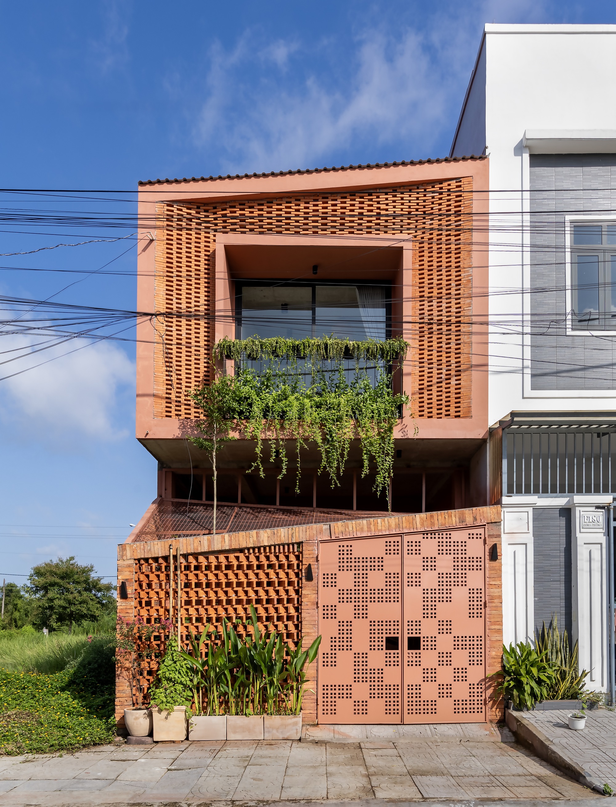 Street facade with perforated brick screen, hanging vines over the balcony and a patterned pink metal gate under a blue sky