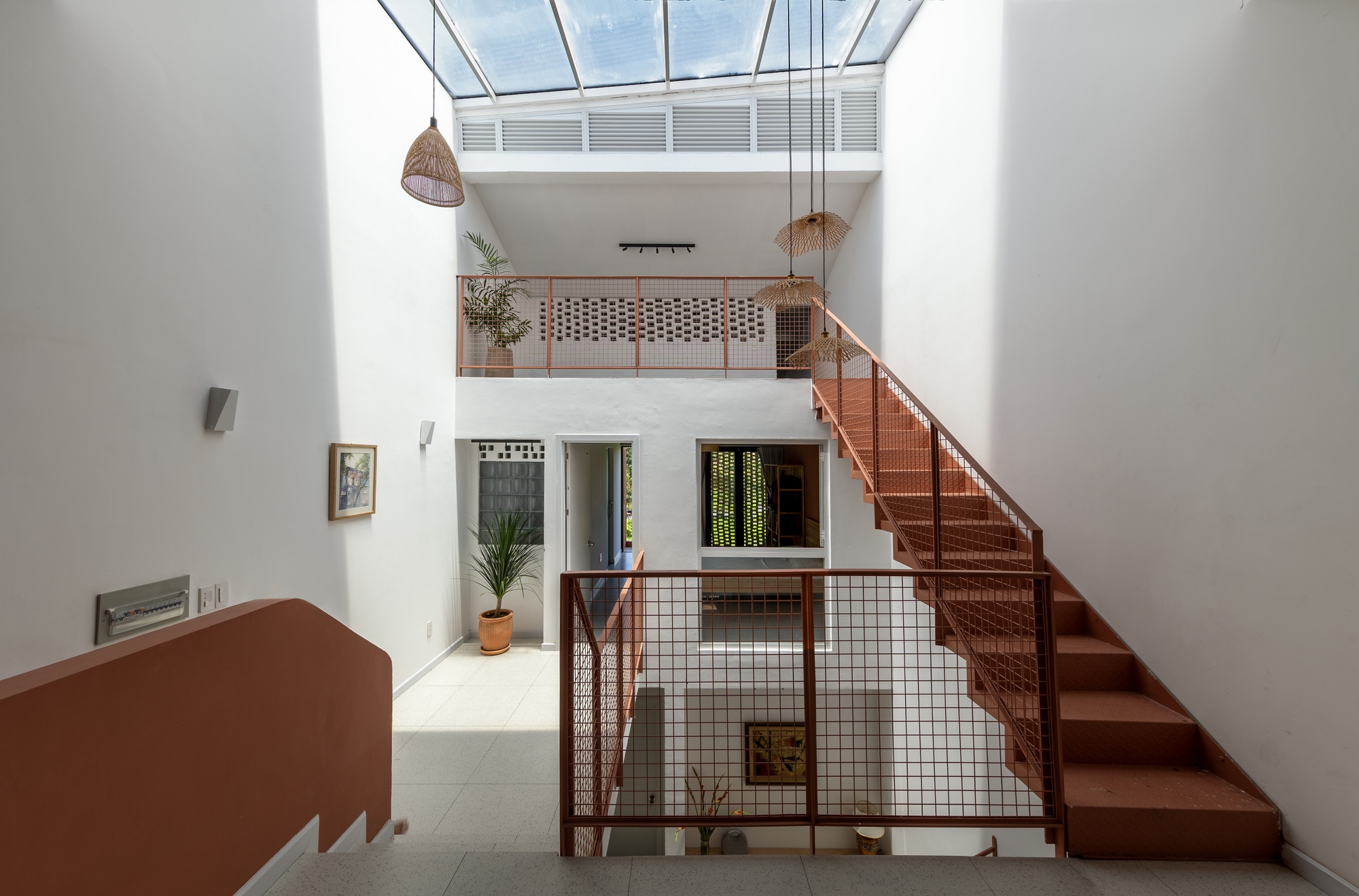 Stairwell under a skylight with a rust-red steel stair, mesh railings and woven pendant lamps in daylight