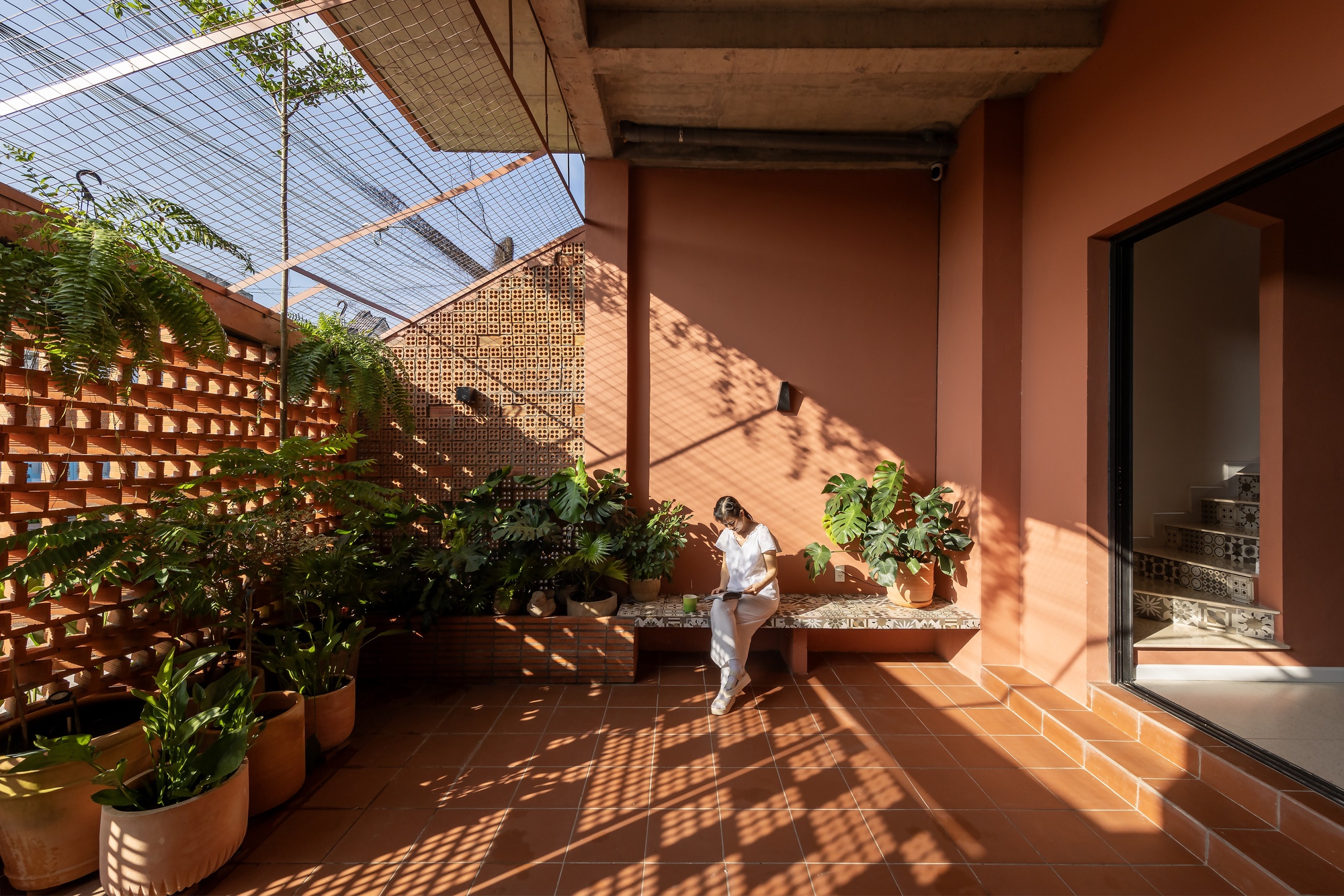 Terracotta-tiled courtyard with a woman seated on a bench among potted plants, sunlight casting lattice shadows from the brick screen