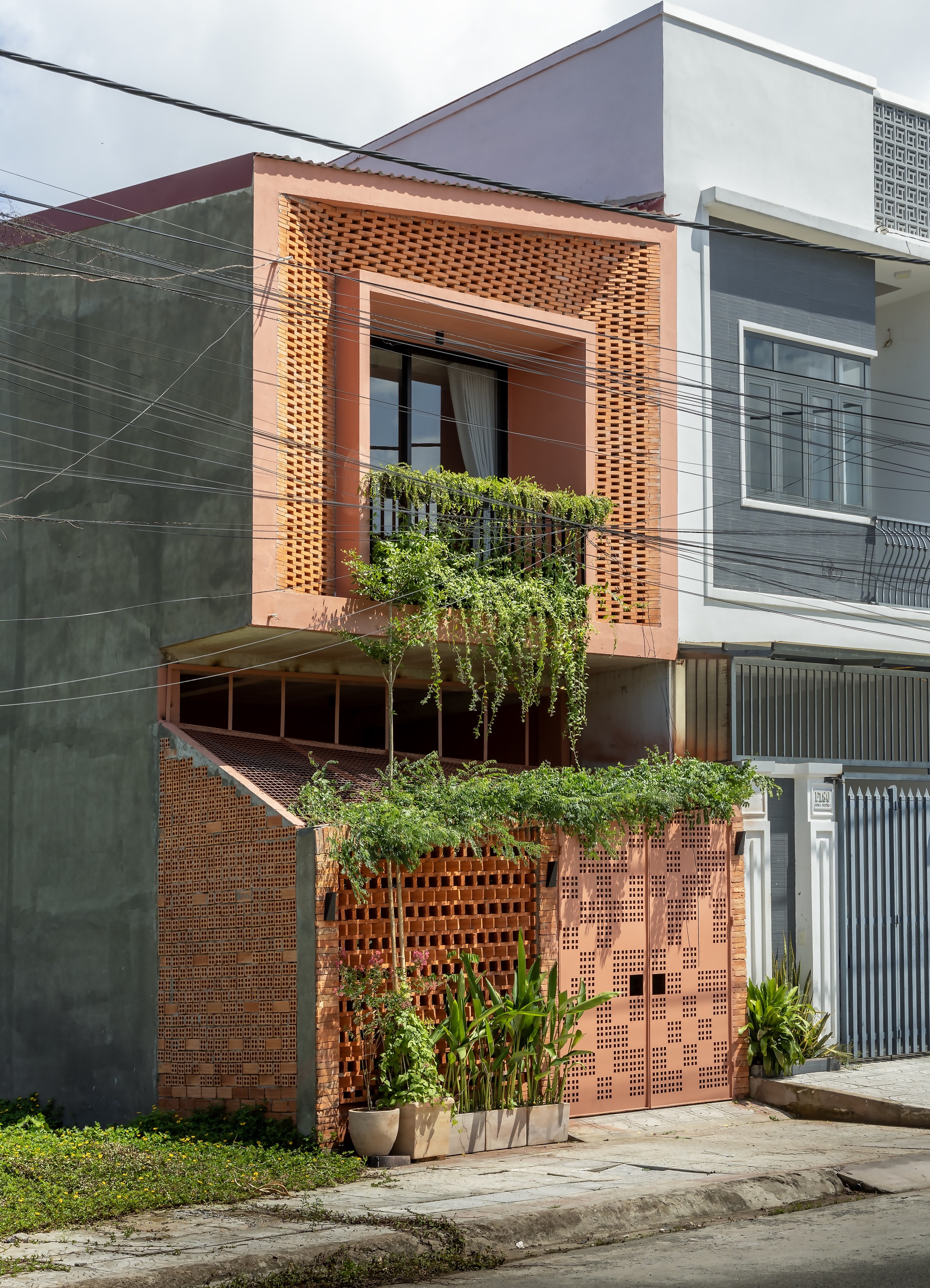 Street facade of terracotta perforated brick and a pierced metal gate, with trailing vines and overhead power lines in daylight