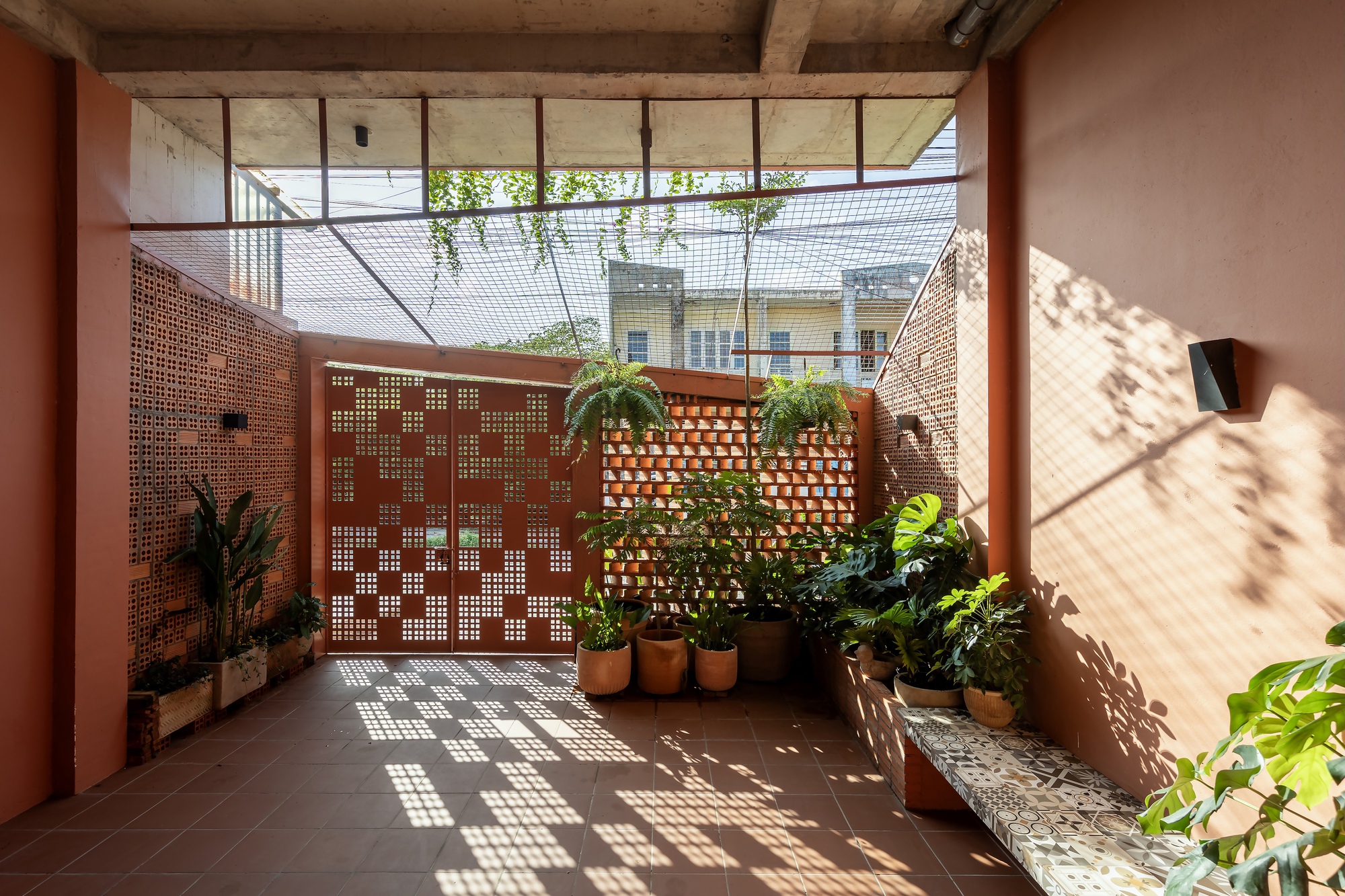 Entry court with perforated gate and brick screens, sunlight casting dappled patterns across the tile floor and a tiled bench