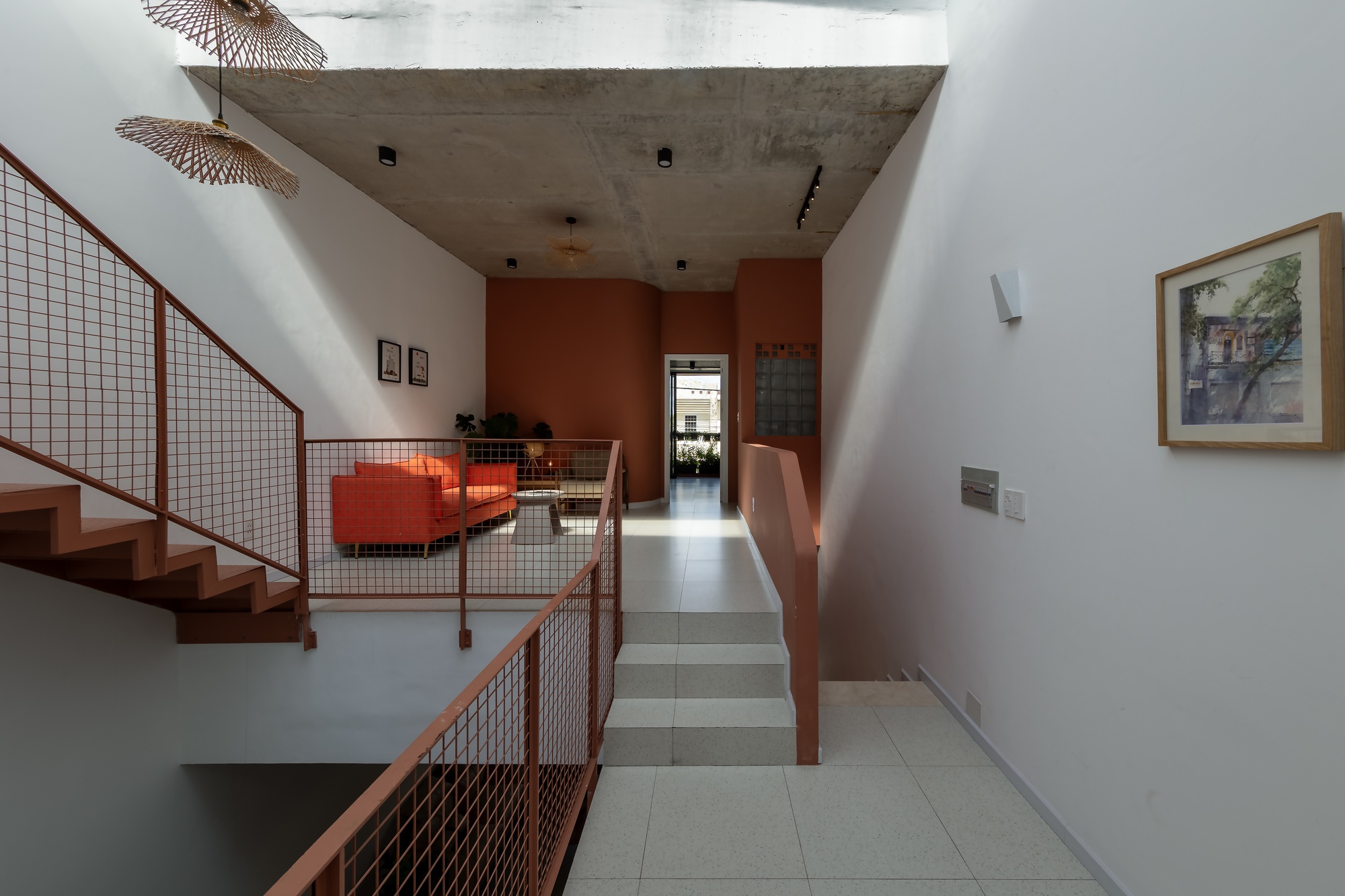 Upper floor corridor with mesh-railed stair, orange sofa in the distance, raw concrete ceiling and a framed painting on the wall