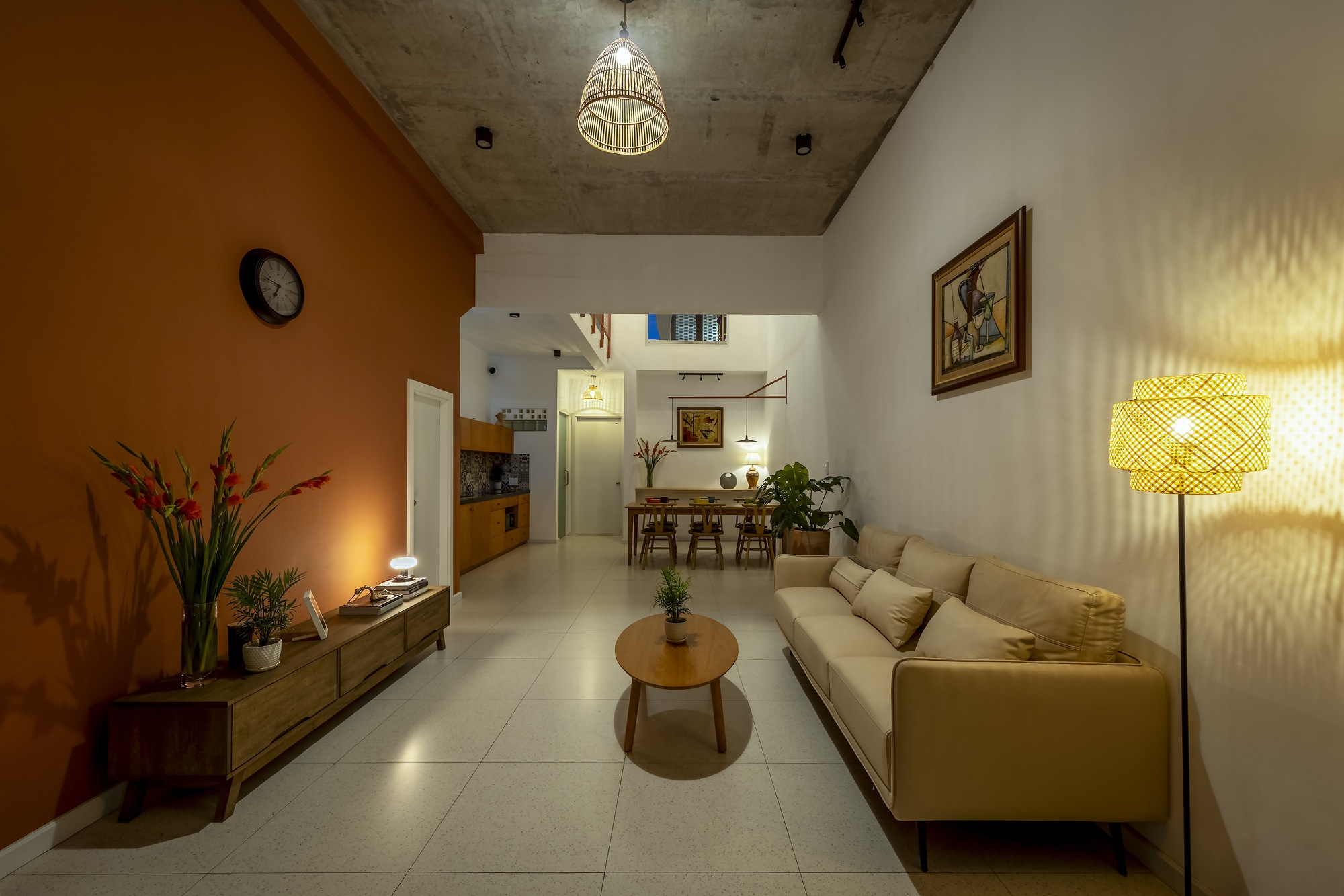 Long living room with exposed concrete ceiling, orange wall and rattan lamps glowing at dusk toward the dining area