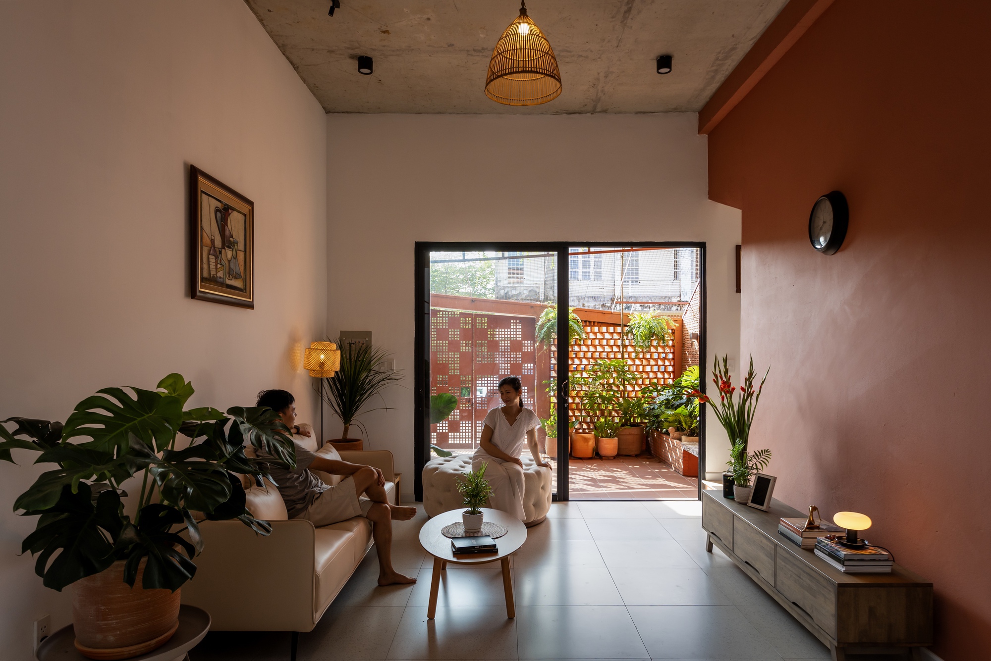 Living room opening through a glass door to a planted terrace with a perforated brick screen, two people relaxing in daylight