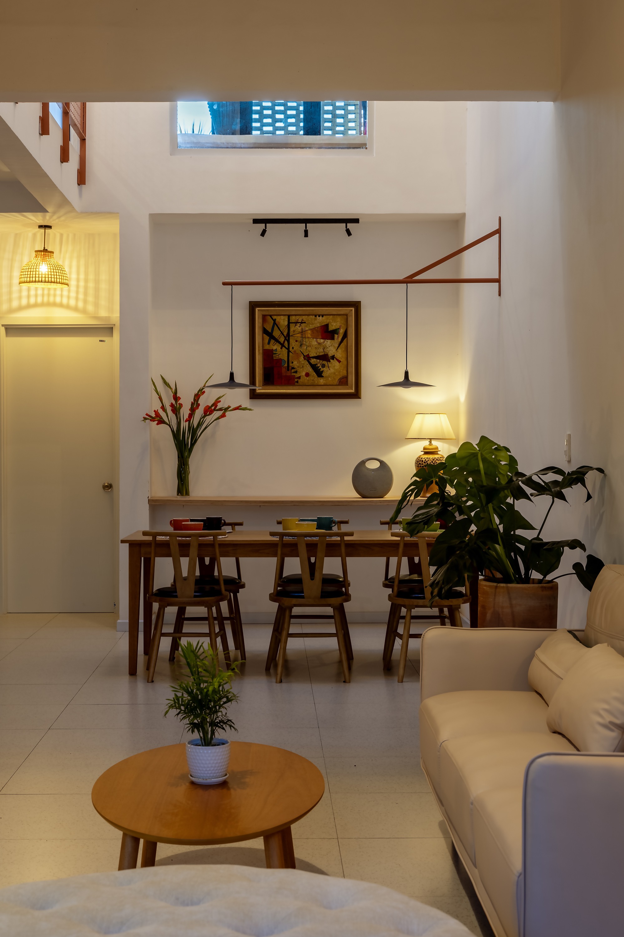 Dining table beneath a framed painting and a copper wall-mounted lamp arm, seen from the sofa in evening light