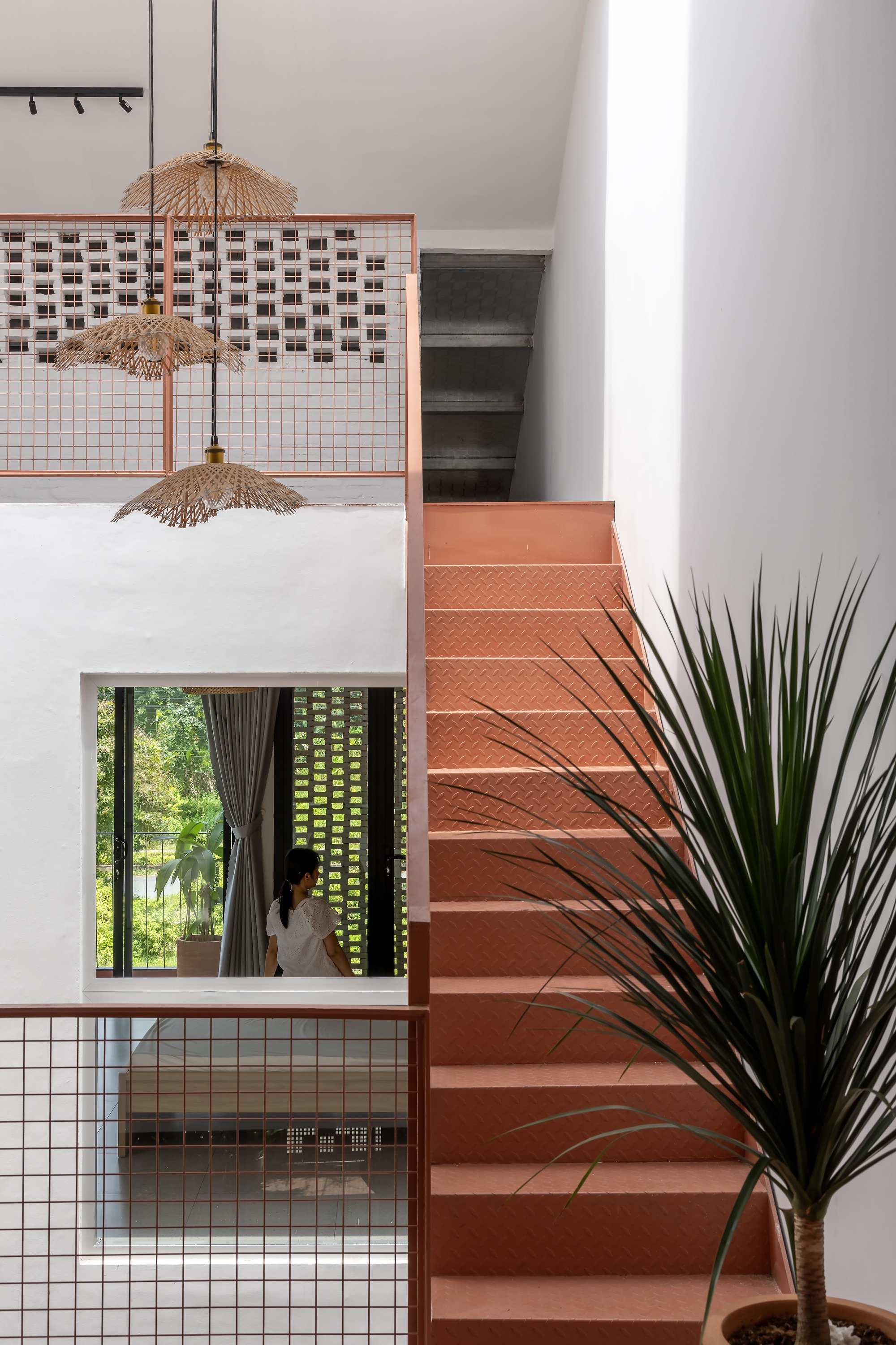 Terracotta-painted stair with textured treads beside a mesh railing, rattan pendants and a woman visible through an interior window