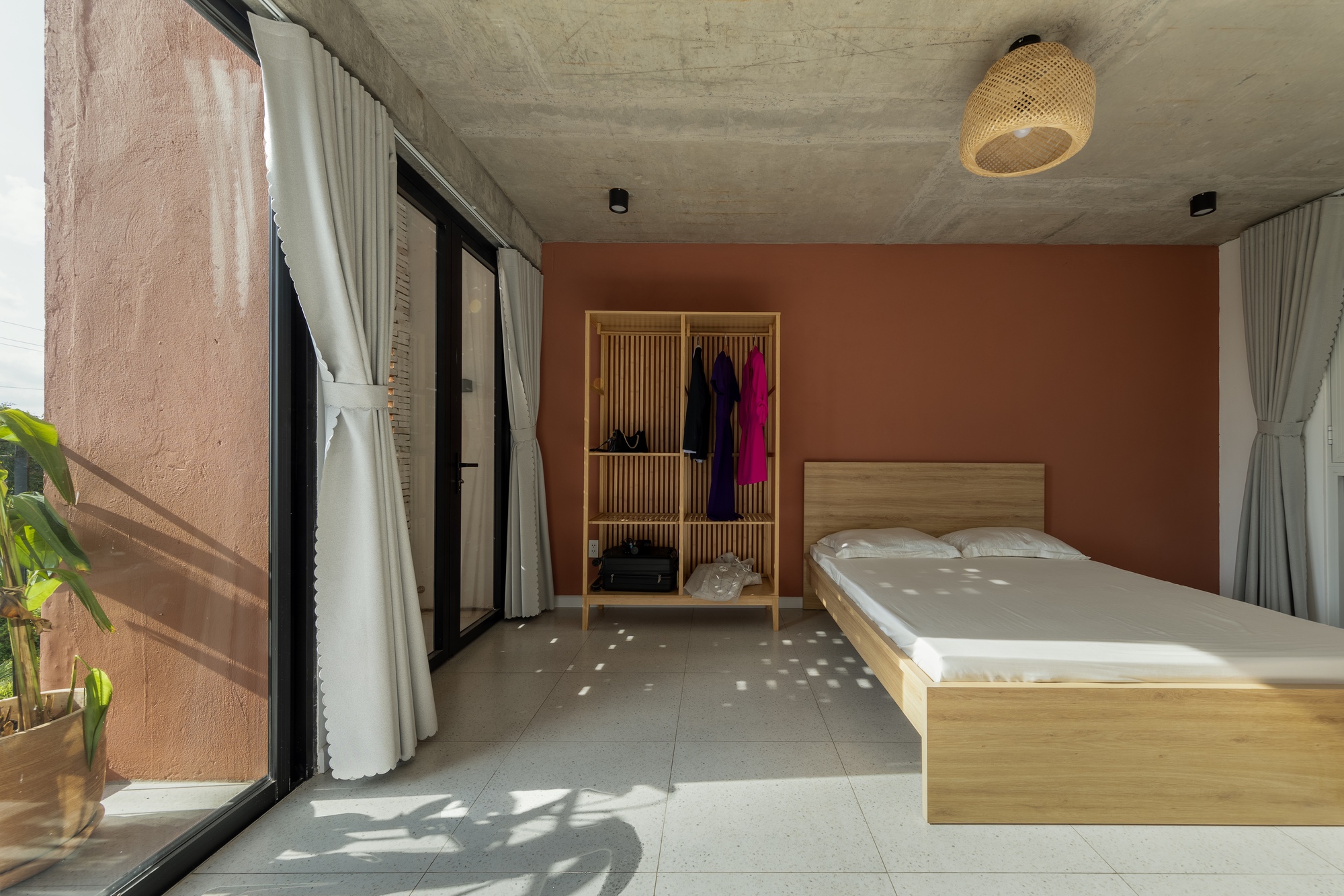 Bedroom with terracotta-painted wall, bamboo wardrobe rack and low timber bed, with dappled sunlight and plant shadows across the terrazzo floor