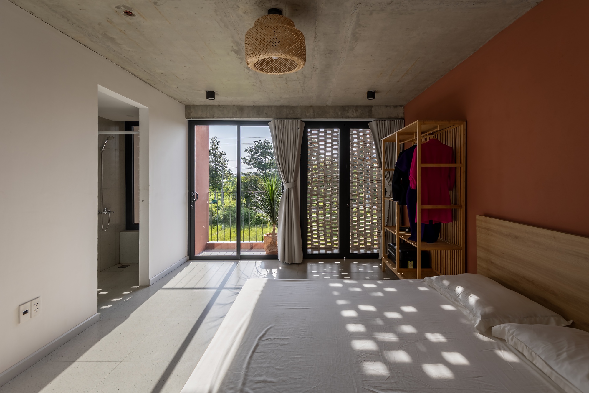 Bedroom looking toward a balcony door and perforated brick screen, with square patches of sunlight falling across the white bedding