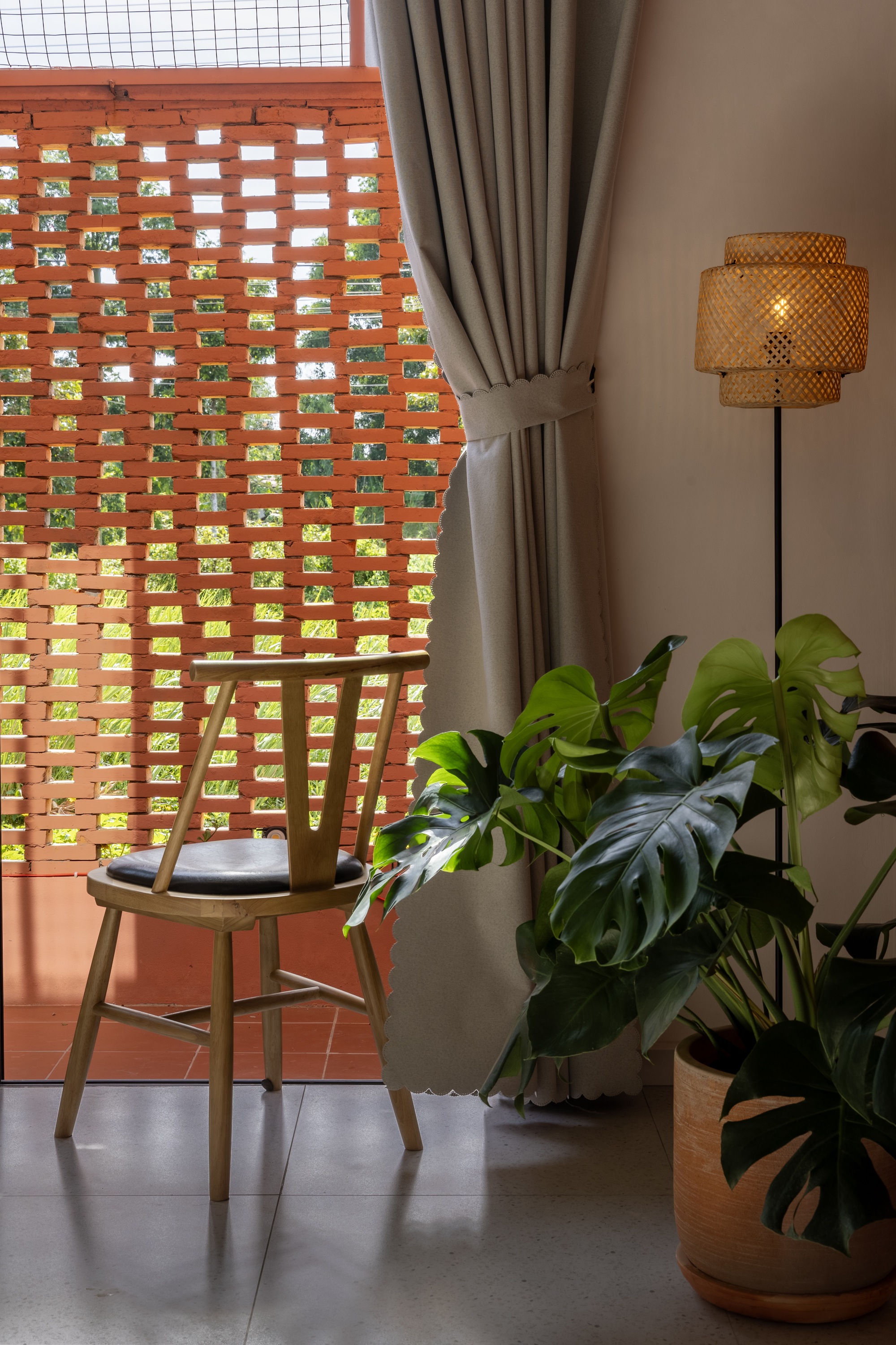 Wooden chair beside a perforated brick screen, with a grey curtain, rattan floor lamp and potted monstera in afternoon light