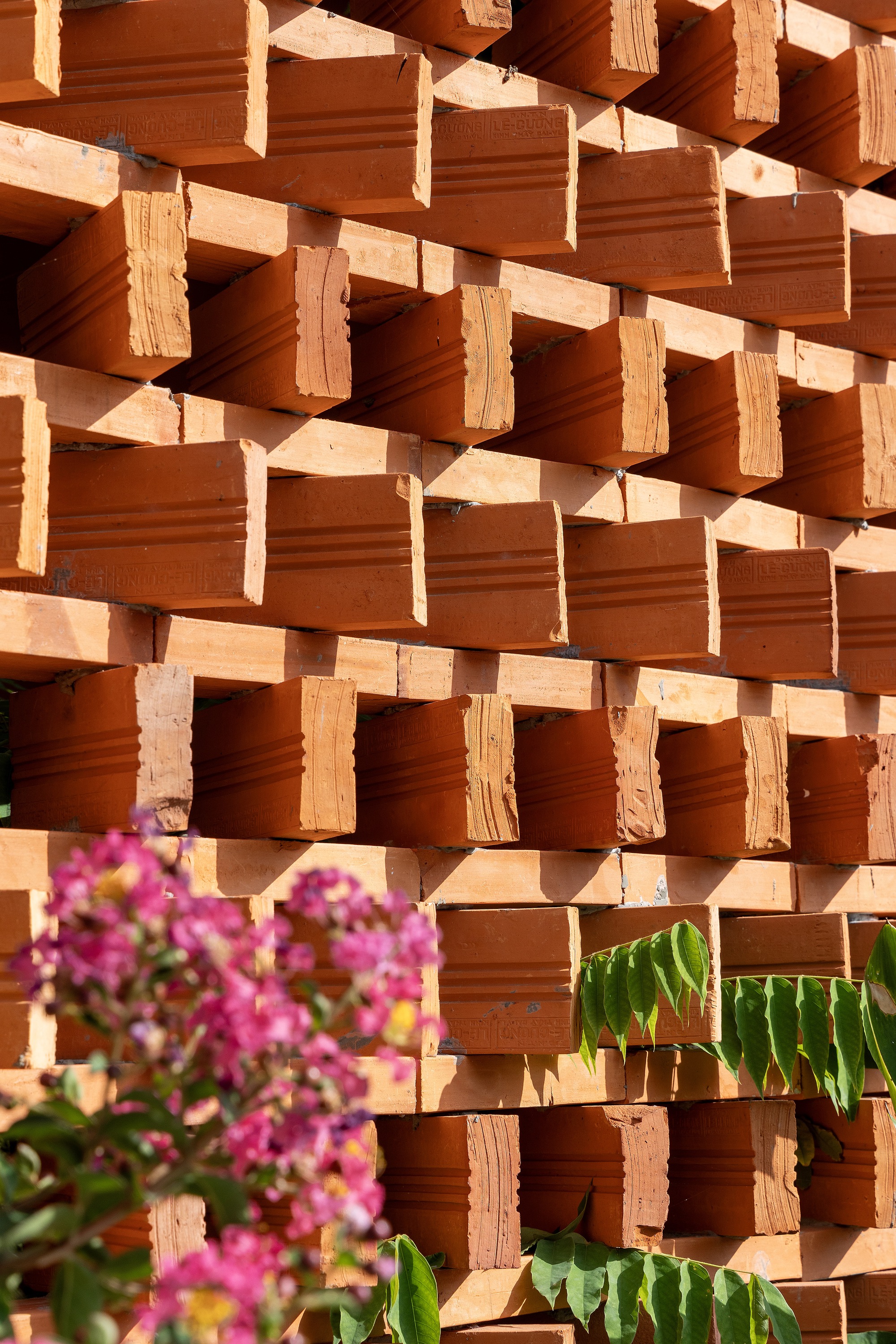 Close-up of stacked terracotta bricks laid with open gaps, with pink flowering branches and green leaves in the foreground