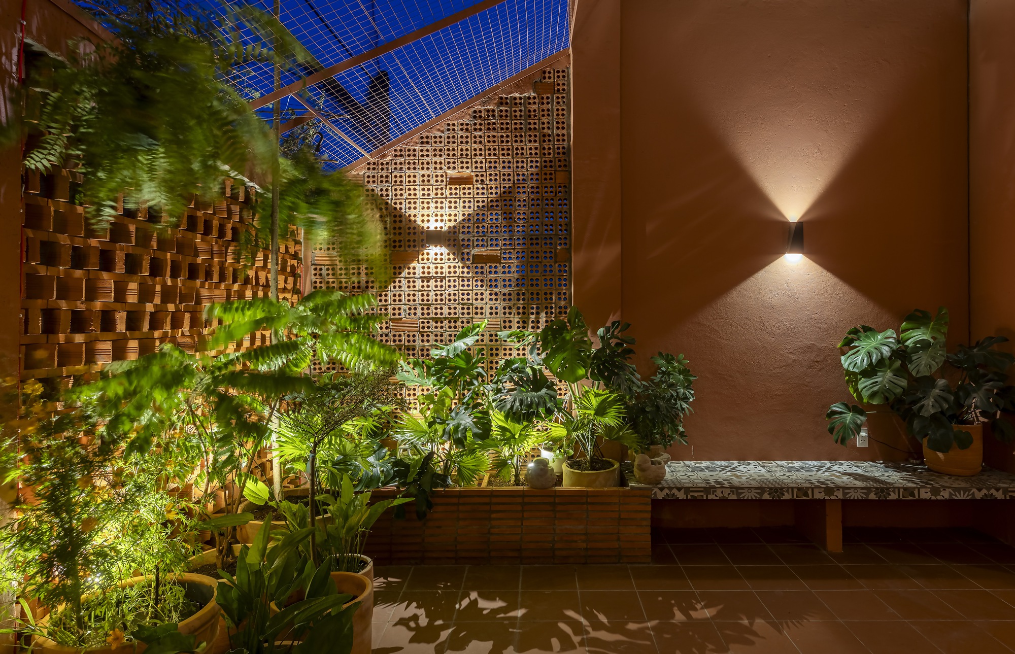 Evening courtyard with a wall light on an orange plaster wall, a tiled bench and ferns beside brick screens under netting
