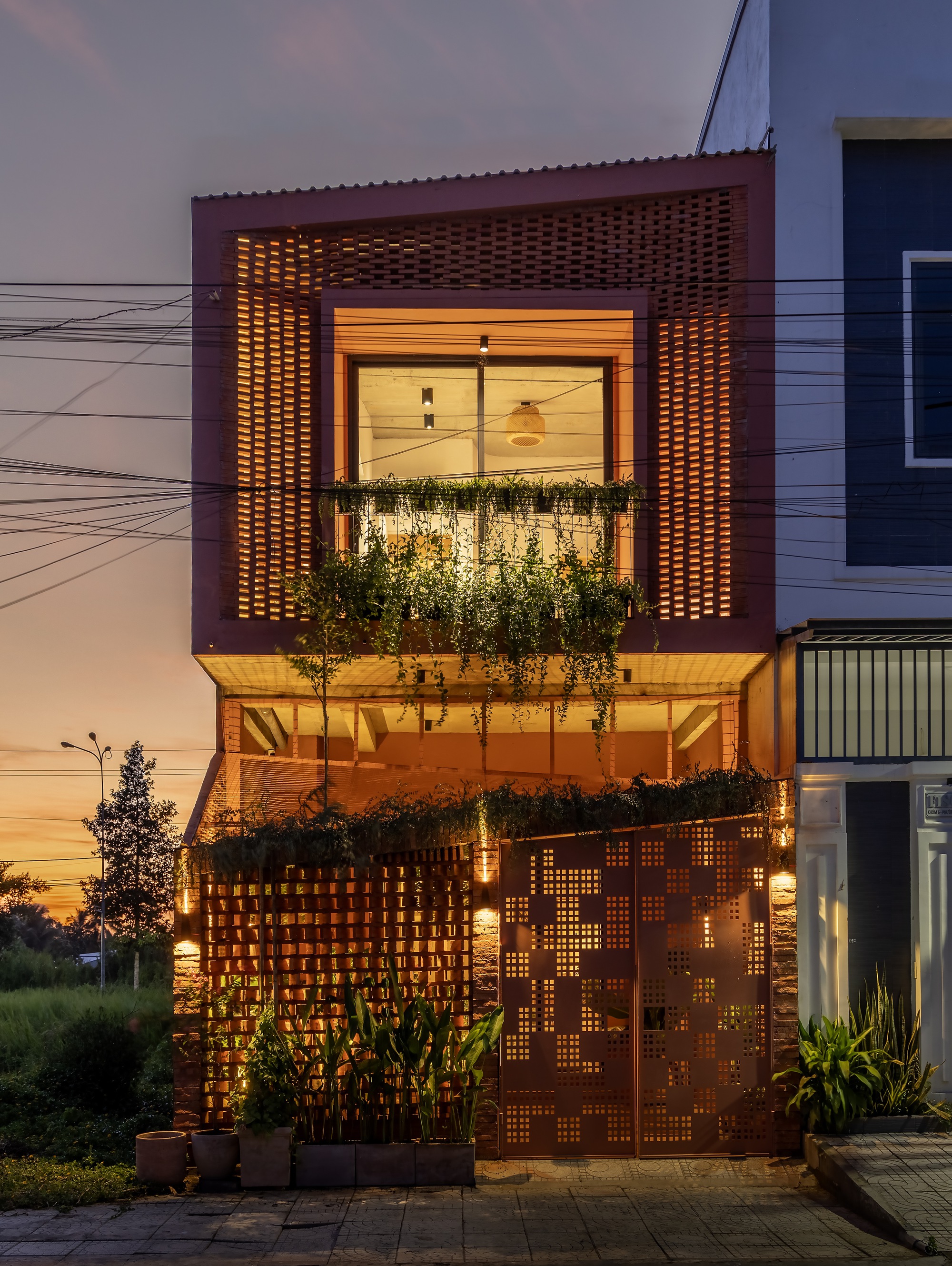 Street facade at dusk with a perforated brick screen above a balcony of hanging plants and a patterned metal gate