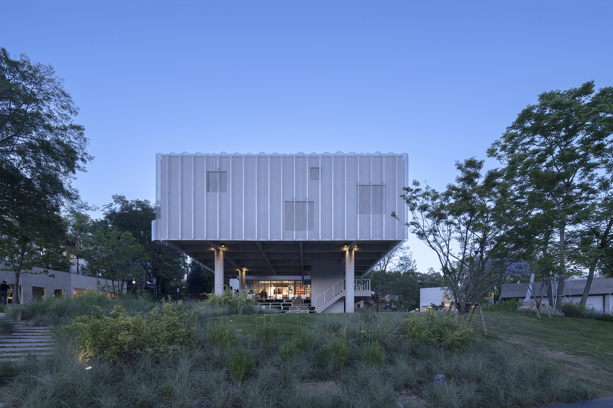 Elevated box clad in white corrugated panels on slender columns, lit from below at dusk above a grassy planted slope