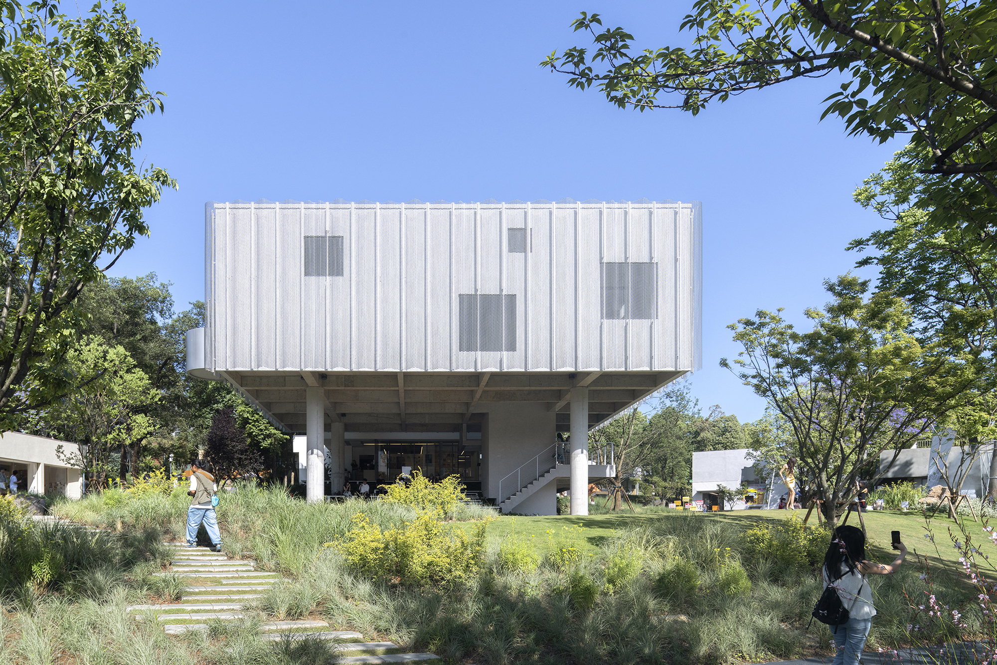 Front view of the raised panel-clad box on columns, with a stone stepping path and visitors in dappled sunlight