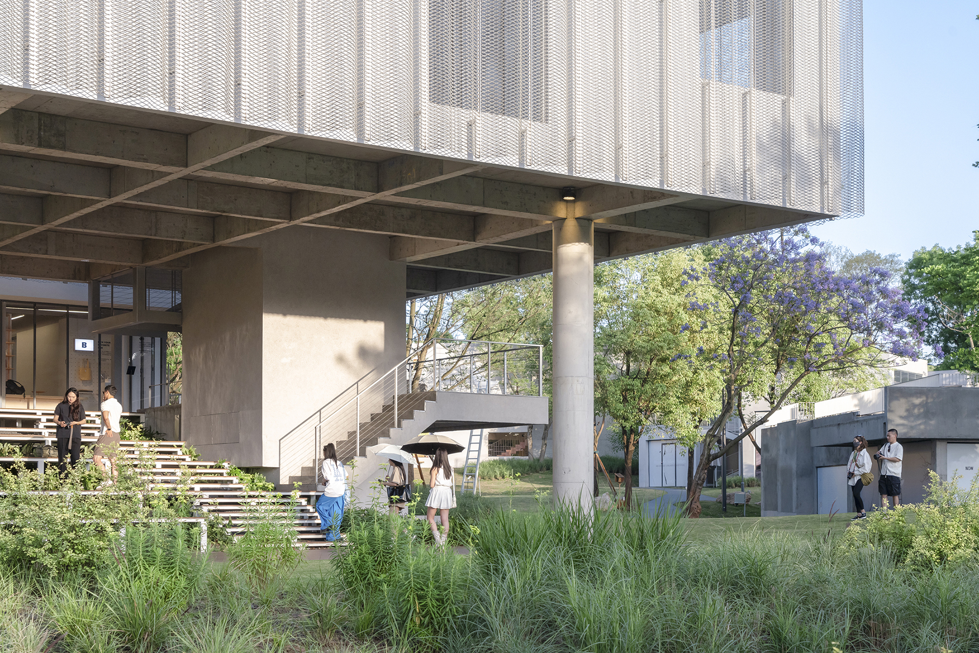 Concrete beams and a round column under the cantilevered mesh-clad volume, with a stair and visitors beside a flowering tree
