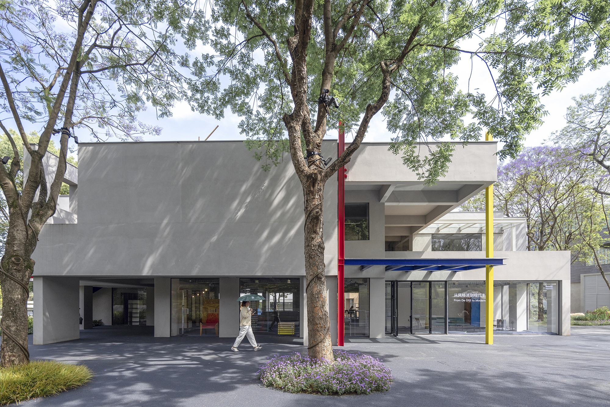 Grey concrete pavilion with red and yellow posts and a blue canopy, a woman with an umbrella walking past a tree