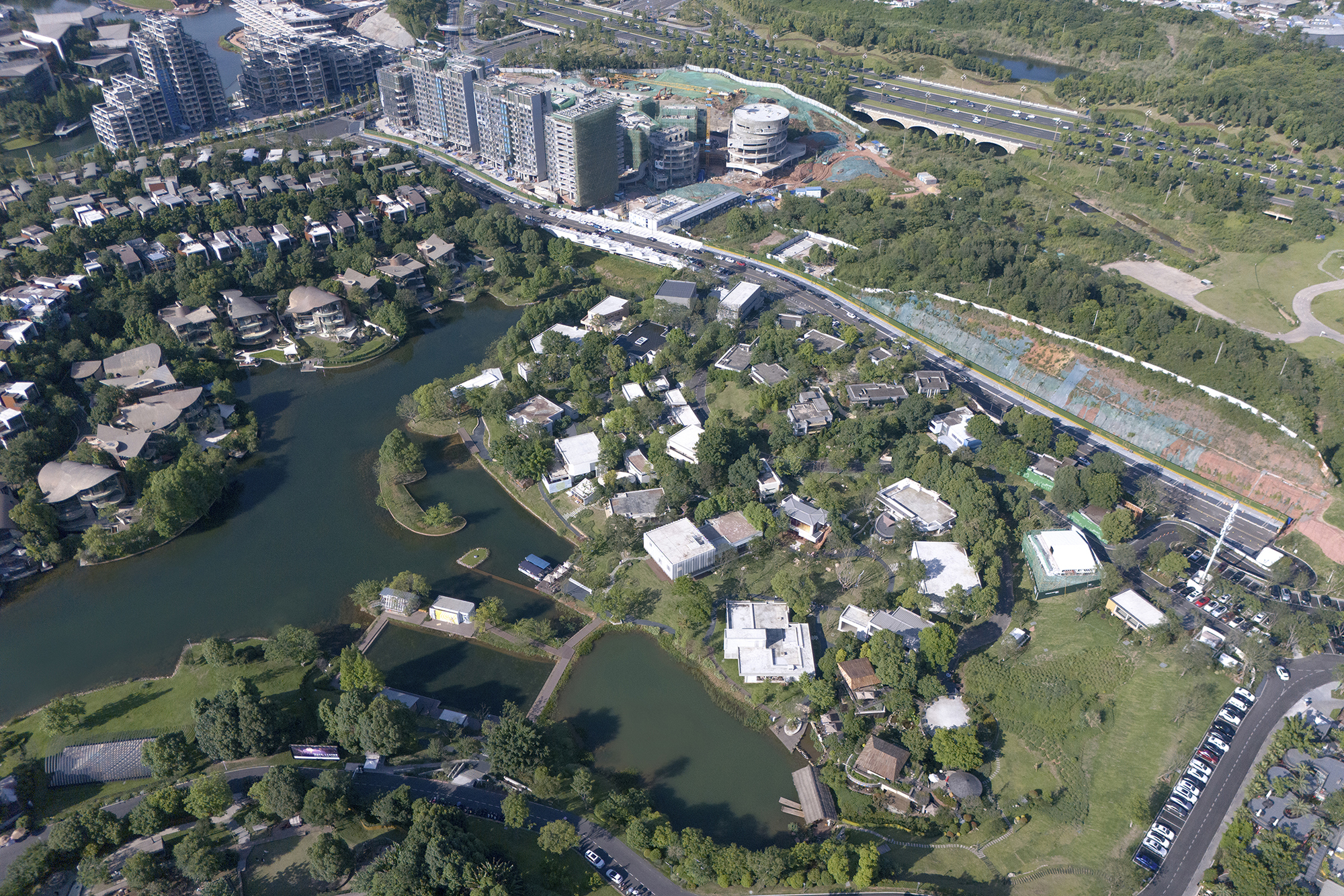 Aerial view of low buildings scattered between lakes and woodland beside a curving road, with apartment towers behind in clear daylight